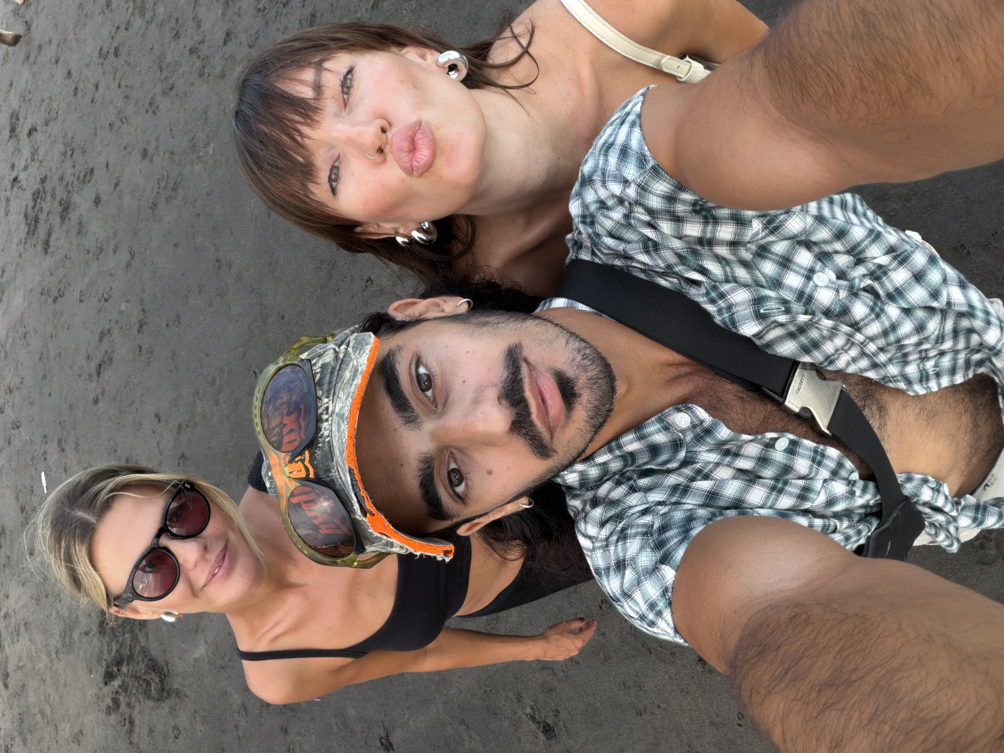 An overhead group selfie captures three individuals outdoors, likely on a dark sand or dirt beach in North Kuta, Indonesia.
In the upper portion of the frame, a woman with brown hair and bangs looks towards the camera, puckering her lips playfully. She wears a light-colored top, large silver hoop earrings, and a nose ring. Adjacent to her, a man lies on the ground, his body oriented towards the left of the image. He has dark hair and a beard, looking directly at the camera with a neutral expression. He is wearing a green and white plaid shirt, a black strap across his chest (with "CALZETTI" visible on the buckle), and a camouflage-patterned hat with orange trim, on which a pair of sunglasses rests. Another woman with blonde hair is positioned towards the bottom left, wearing sunglasses and a black top, offering a faint smile towards the camera.
The setting appears to be a natural, outdoor environment under clear daylight. The ground is dark and textured, consistent with sand or volcanic earth. The overall mood is casual and relaxed, indicative of friends enjoying a moment together in a tropical location.
