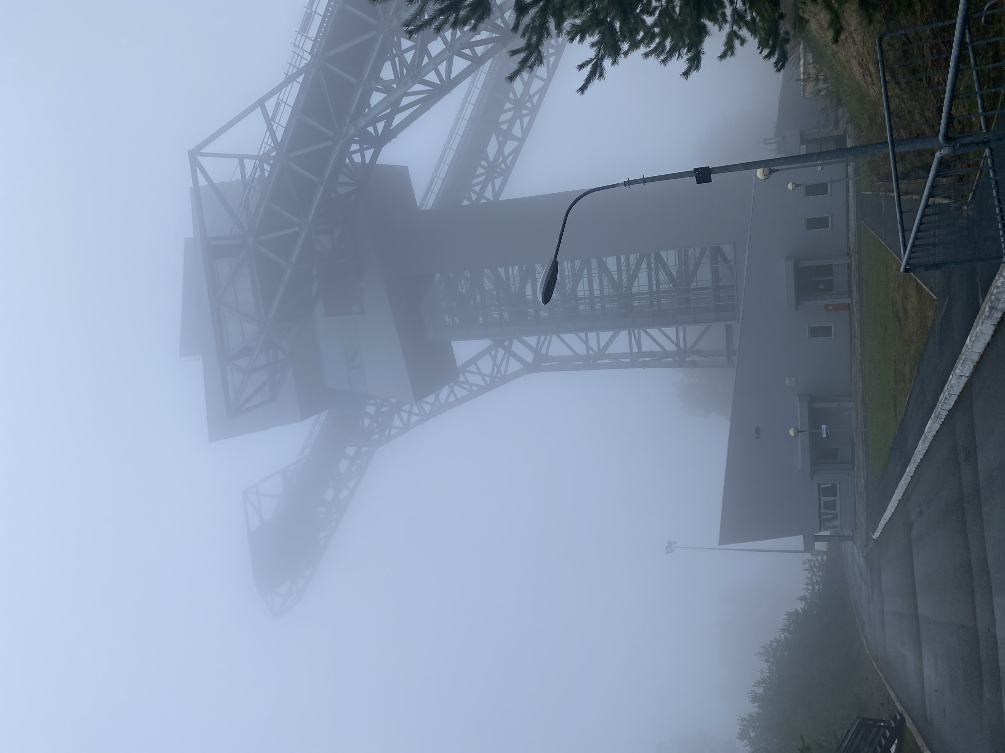 The image depicts a large metal structure resembling a gantry crane shrouded in heavy fog. It is located next to a gray, rectangular building with several visible windows and a door. A street lamp is positioned between the structure and the building. A section of paved roadway runs along the side of the building, with a small patch of green vegetation visible next to the road. The heavy fog obscures the background and other details. Based on the presence of the structure and the location provided, the scene appears to be an industrial area in Прикамский, Russia.