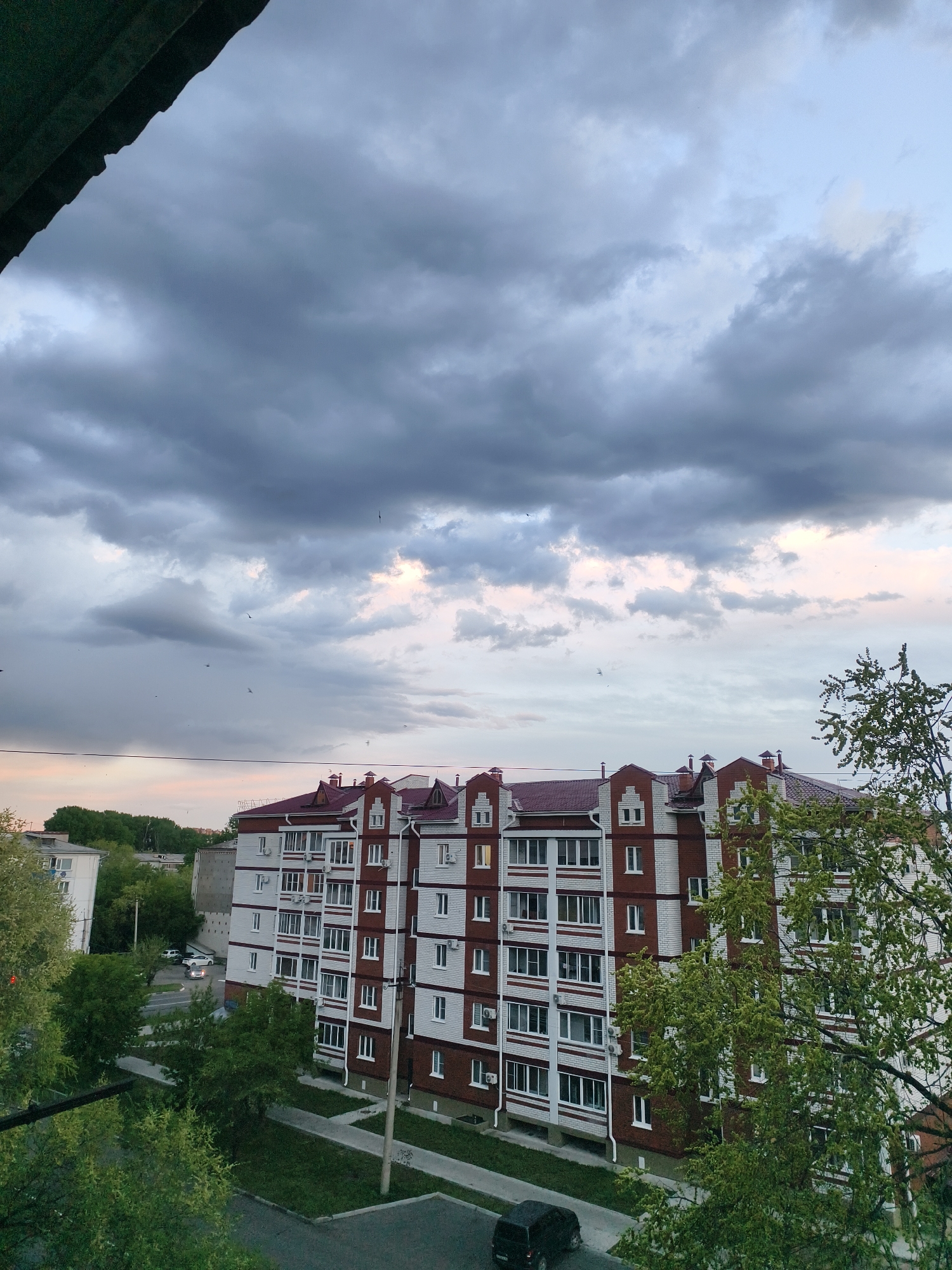 The image shows a view in Belogorsk, Russia. A large, multi-story apartment building dominates the scene, constructed from red brick and white panels, topped with a red roof. The building is surrounded by trees, some partially obscuring the view. A street runs alongside the building, with a dark-colored vehicle parked. The sky is overcast with dark, layered clouds, hinting at a possible impending storm or fading daylight, with a narrow strip of light visible along the horizon. A portion of a roof overhang is visible in the upper left corner, indicating the perspective is from an elevated position. Small birds can be seen flying in the distance.