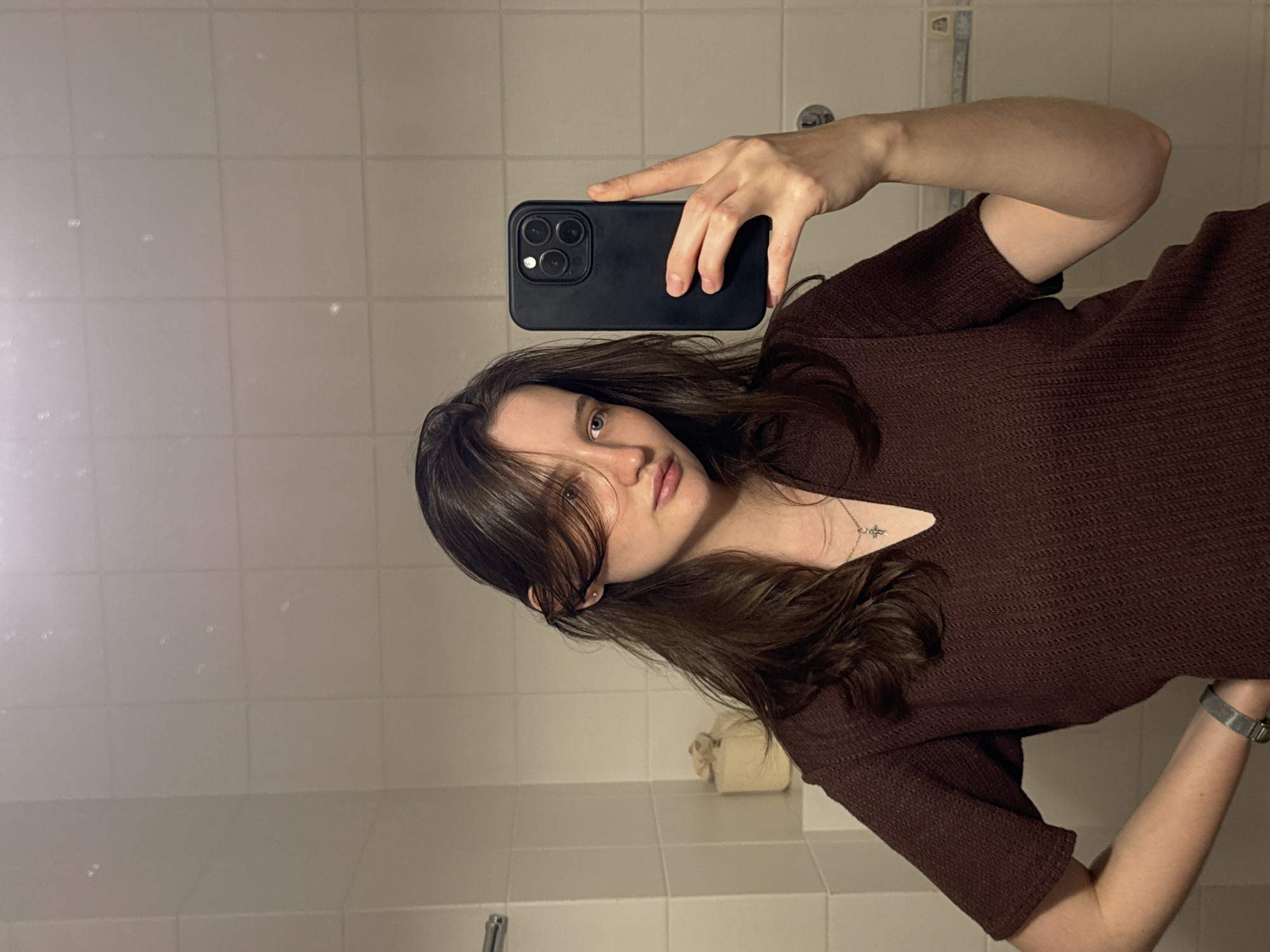 A young woman with long, dark brown hair is taking a selfie in a bathroom. She is wearing a dark brown, short-sleeved shirt and a silver necklace with a cross pendant. Her left hand is on her hip, and her right hand is holding a black smartphone, positioned to capture her reflection in the mirror. The bathroom features white tiled walls and a tiled floor. A roll of toilet paper is visible on a shelf to the left of the woman. The lighting appears to be artificial. The specific location of Журавлево (кемеровский), Russia, cannot be determined from the visual information provided in the image.
