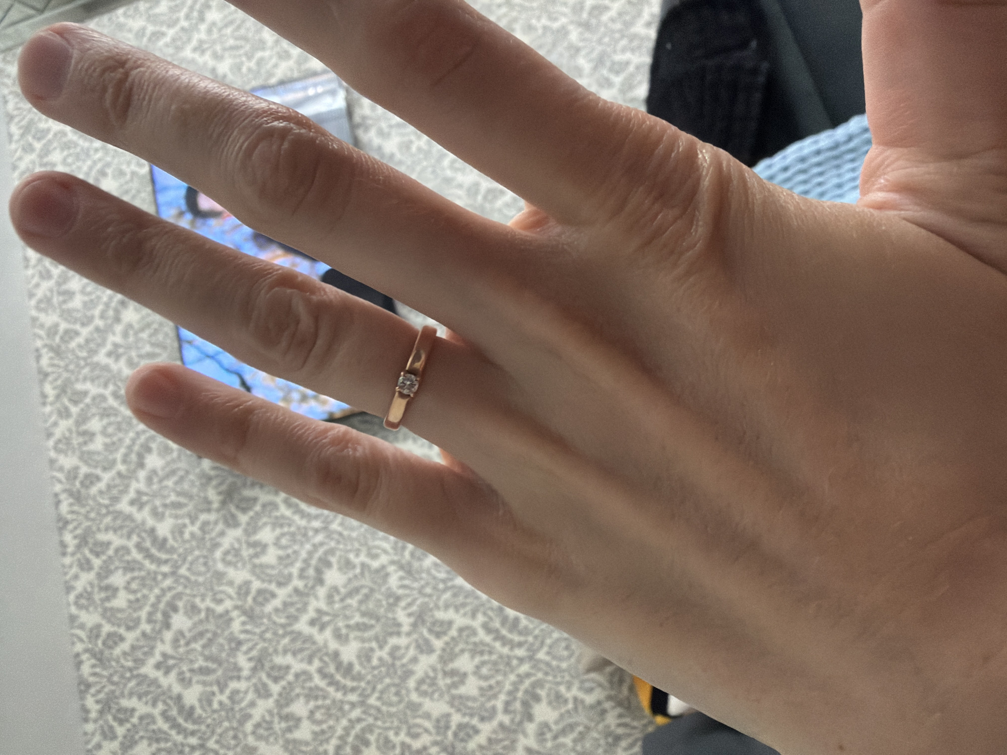 An overhead, eye-level, medium shot shows a woman's left hand with a rose gold band and a small diamond ring on her ring finger. Her hand is positioned in the foreground and slightly angled to the right, with her fingers extended. The background features a patterned wallpaper in shades of gray and white, and a blurred television screen displaying a person. The scene is indoors, likely in a residential setting. There are no discernible details about the location in Bila Tserkva, Ukraine.