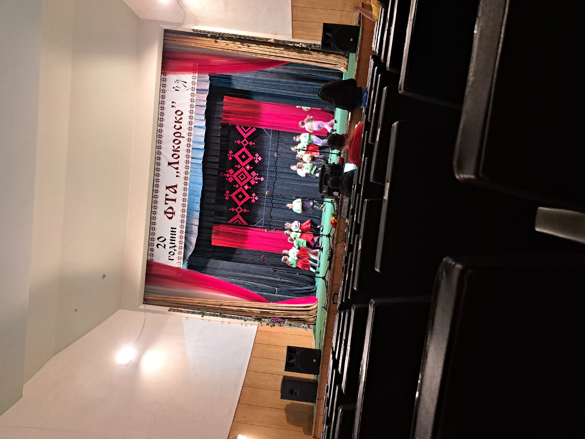 The image captures an indoor performance taking place in an auditorium or cultural hall, seen from the perspective of an audience member in the front row. The main subject is a brightly lit stage where several children, likely performers, are standing. They are dressed in colorful outfits, some in red and green, others in white and green tops with red skirts or pants, possibly traditional folk costumes. Each child appears to be holding a microphone, suggesting they are either singing or speaking. An adult figure is partially visible on the right side of the stage.
The stage is set with a backdrop featuring red and blue curtains, with a central black panel adorned with a large red and black geometric pattern, resembling traditional embroidery or weaving. Above the stage, a white banner with text is displayed. The text, in Cyrillic, reads "20 години ФТА „Локорско”". This translates to "20 years FTA "Lokorsko"", indicating a celebration of the 20th anniversary of a Folk Dance Ensemble (FTA) from Lokorsko, Bulgaria.
The foreground shows rows of dark, empty audience seats, suggesting the photo was taken before or during a performance with limited attendance, or from a specific vantage point. Two large black speakers are placed on the floor to the left of the stage. The hall's ceiling and upper walls are visible, illuminated by bright overhead lights. The overall scene depicts a cultural event or celebration in Lokorsko, Bulgaria. The image is rotated 90 degrees clockwise.