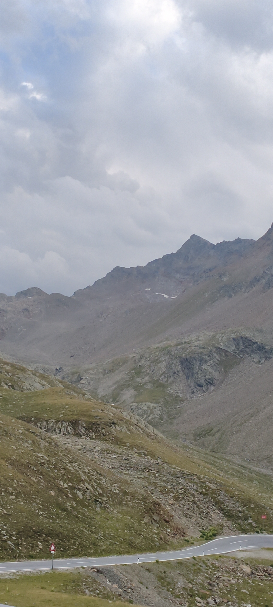 The image displays a cloudy, overcast sky above a rugged, mountainous landscape. The mountains are characterized by steep slopes, rocky terrain, and patches of sparse vegetation. A winding road with white lines marking the lanes traverses the lower portion of the image, bordered by a grassy embankment and rocky scree. A red and white diamond-shaped traffic sign is visible on the left side of the road. The scene appears to be a high-altitude road in a natural, undeveloped area, possibly in the Austrian Alps, given the mention of Sölden, Austria, which is a well-known ski resort town located in the Ötztal valley, surrounded by high mountain peaks. There are no people or animals visible in the image.