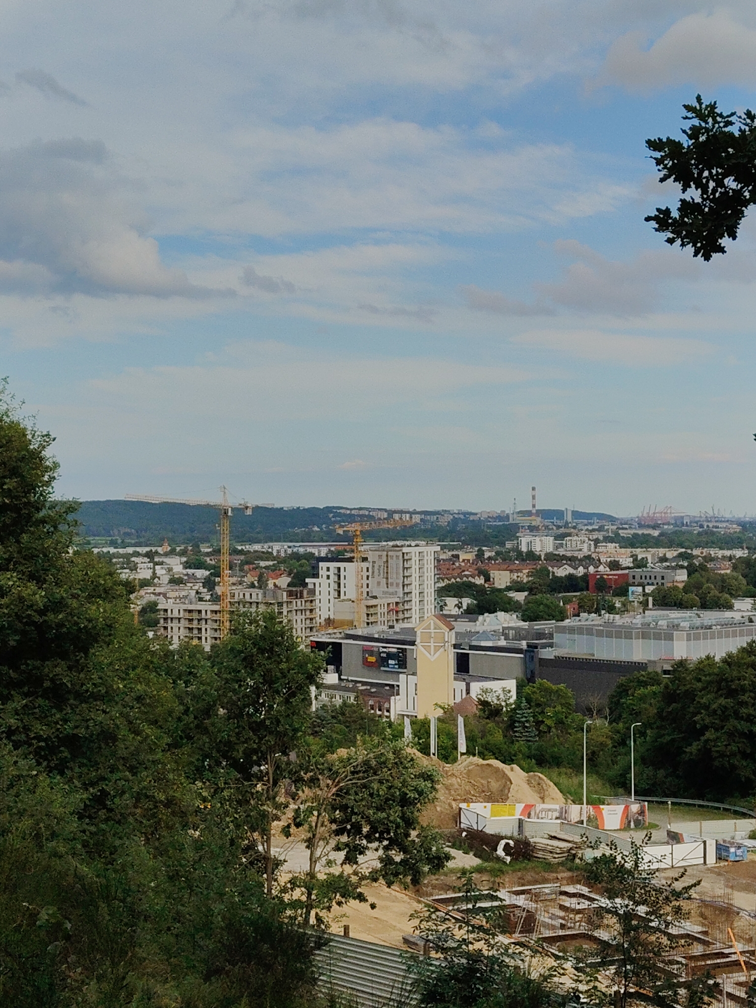 A panoramic view of a city landscape in Rumia, Poland, is captured from an elevated perspective, looking out over a developing urban area. In the foreground, lush green trees obscure a portion of the scene. Beyond the trees, a construction site is visible, with the foundations of several buildings laid out and scaffolding in place, suggesting ongoing development. Further into the cityscape, a cluster of modern buildings, including apartment complexes and commercial structures, dominates the middle ground. Several construction cranes are dotted throughout this area, further emphasizing the active construction. In the background, a hazy horizon stretches across the frame, featuring distant buildings, industrial structures, and a prominent smokestack with a red and white top. The sky above is partly cloudy, with scattered white clouds against a pale blue backdrop.