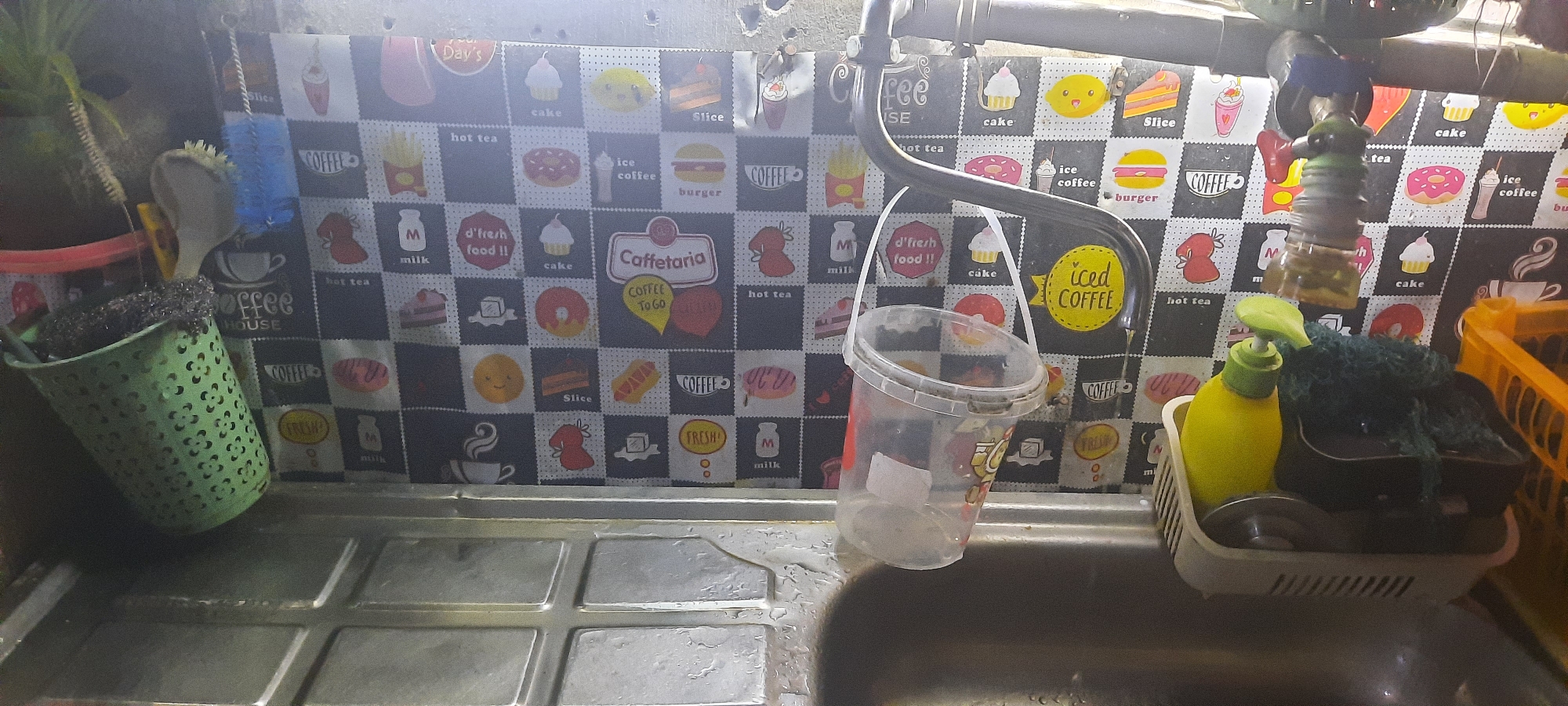 The image depicts a close-up view of a kitchen sink area. The background features a black and white tiled wallpaper with various food and beverage graphics, including cupcakes, donuts, burgers, and coffee cups. A chrome faucet with a red handle is visible, with water dripping from it. To the left of the faucet, a clear plastic bucket with a white lid and a white string handle is hanging. Next to the bucket, a green perforated plastic container holds a brush. To the right of the faucet, a yellow soap dispenser with a green nozzle is placed in a white plastic dish rack, which also contains a dark green scrub pad and a black sponge. In the background, a yellow plastic basket is partially visible. The sink itself is made of stainless steel and appears to be wet. The overall impression is that of a functional, albeit somewhat cluttered, kitchen workspace. The location in Jatitengah, Indonesia, is provided as context for this scene.