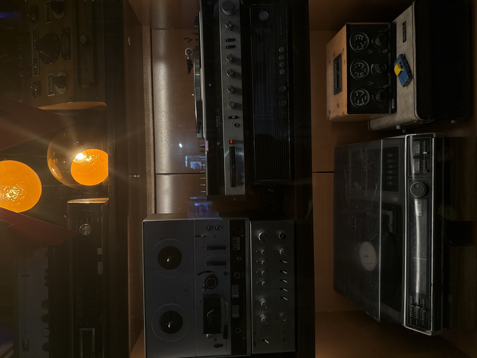 A display cabinet showcases vintage audio equipment. A reel-to-reel tape recorder sits beside a control panel with numerous knobs. Above are stacked components: a Sony unit with a cassette deck and a tube radio with gauges. A small toy car rests on top of one unit. Light fixtures with glowing orbs are visible near the equipment. The setting is dim, with reflective surfaces adding to the visual complexity.