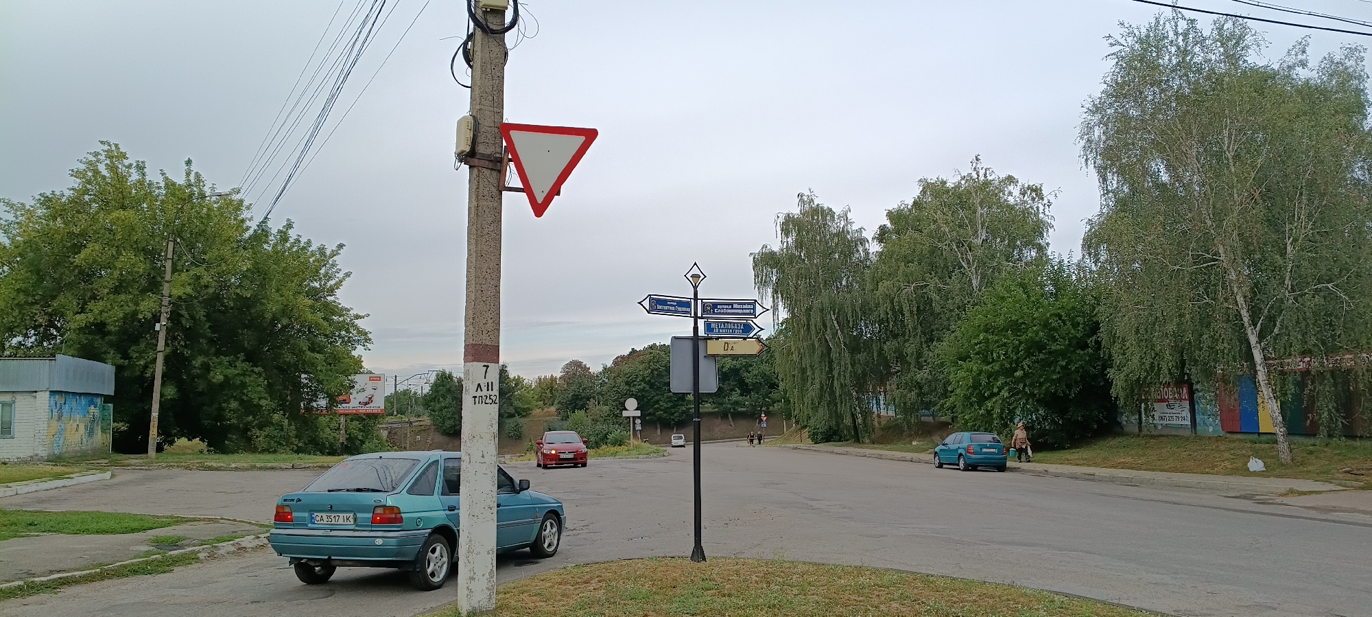 The image depicts a street scene in Kamianka, Ukraine, on an overcast day. In the foreground, a teal-colored hatchback car is parked facing left, with its rear to the camera. To the left of the car, a utility pole stands prominently, displaying a triangular yield sign and some handwritten numbers and letters. Further down the road, a red sedan is visible.
A distinctive directional signpost stands near the utility pole, featuring multiple arrows indicating directions with place names in Ukrainian, such as "Антоніна Гужва" (Antonina Guzhva), "ПУТЕВАЯ КАЗАЧКА" (Putevaya Kazachka), "МЕТАЛЛБАЗА" (Metalbaza), and "д.А" (d.A).
In the background, several trees with green foliage line the street. A small, light-colored building with blue and yellow accents is situated on the left side of the image. On the right side of the road, another teal-colored car is parked, and a person is walking on the sidewalk. A billboard with text and images of garden equipment is visible in the distance on the right. The overall environment suggests a residential or mixed-use area within the city.