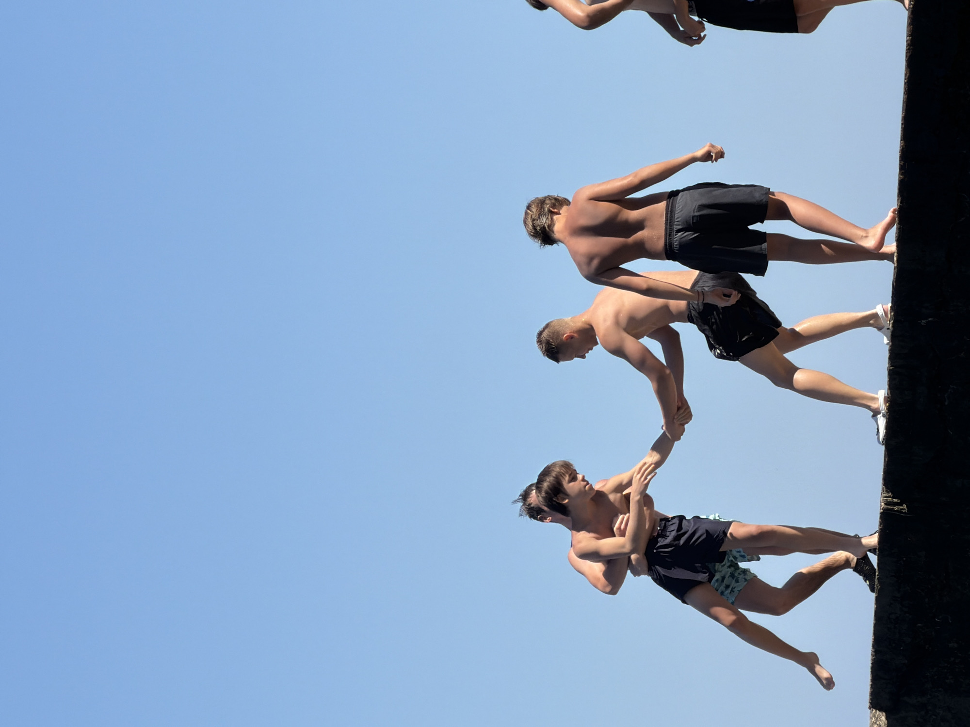 On a sunny day in Partenit, Ukraine, five young men stand on a concrete structure with a clear blue sky as the backdrop. Two individuals on the left are engaged in a wrestling match, their bodies intertwined as they balance precariously. Another young man stands to their right, facing away from the camera with his back to the viewer. Further to the right, a fourth young man is also seen from behind, and a fifth individual is partially visible on the far right of the frame. All individuals are wearing swim trunks, suggesting they may be near a body of water or participating in water-related activities. The concrete structure appears to be a sea wall or pier, indicating a coastal or waterfront location.