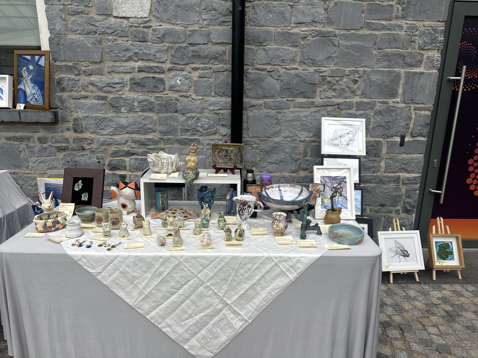 The image depicts an outdoor stall in Kilkenny, Ireland, likely during a market or fair. A grey linen tablecloth drapes a table displaying an assortment of ceramic art pieces, small sculptures, and framed artwork. The background consists of a textured grey stone wall.
On the left, a framed painting of a woman with blue accents is propped on a window ledge. The main table is laden with various ceramic items, including animal figurines (cats, rabbits, dogs), decorative bowls, vases, and whimsical creations. Several small, colorful charms or pendants hang from some of the ceramic pieces.
To the right of the main display, a separate easel holds a framed drawing of a fly. Next to it, another small easel displays a framed painting of an abstract landscape or island-like shape. A dark grey door with a silver handle and a partially visible graphic design is to the far right. The ground is paved with cobblestones. No people are visible in the image.