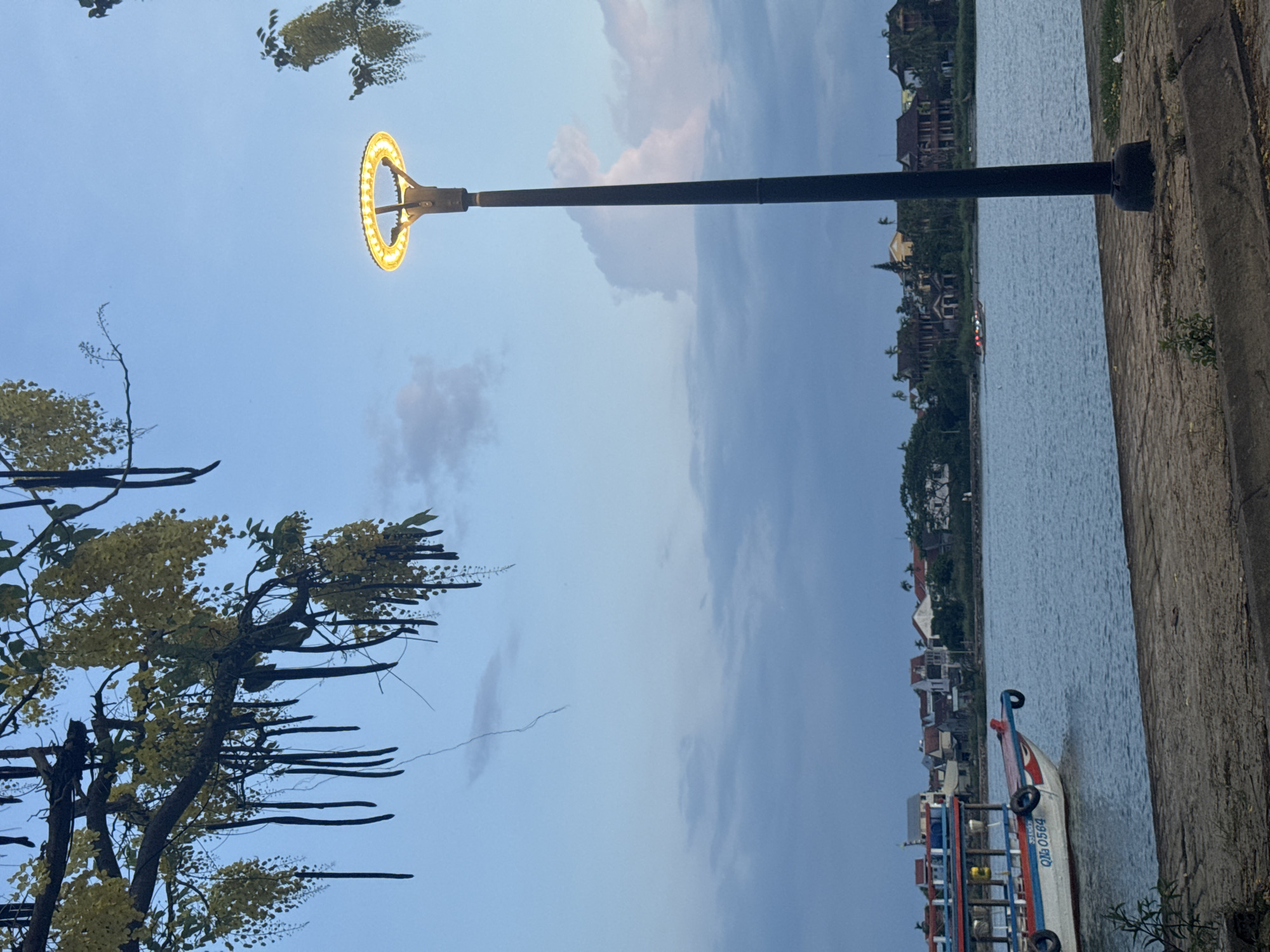 The image shows a riverside scene in Hội An Ward, Vietnam. A black lamppost with a circular, glowing light fixture is positioned along a stone embankment. Across the water are various buildings with diverse architectural styles. A tree with yellow blossoms and long, dark seed pods is partially visible on the left. A small boat with the markings "Qna 0564" is docked along the embankment. The sky is blue with scattered clouds.