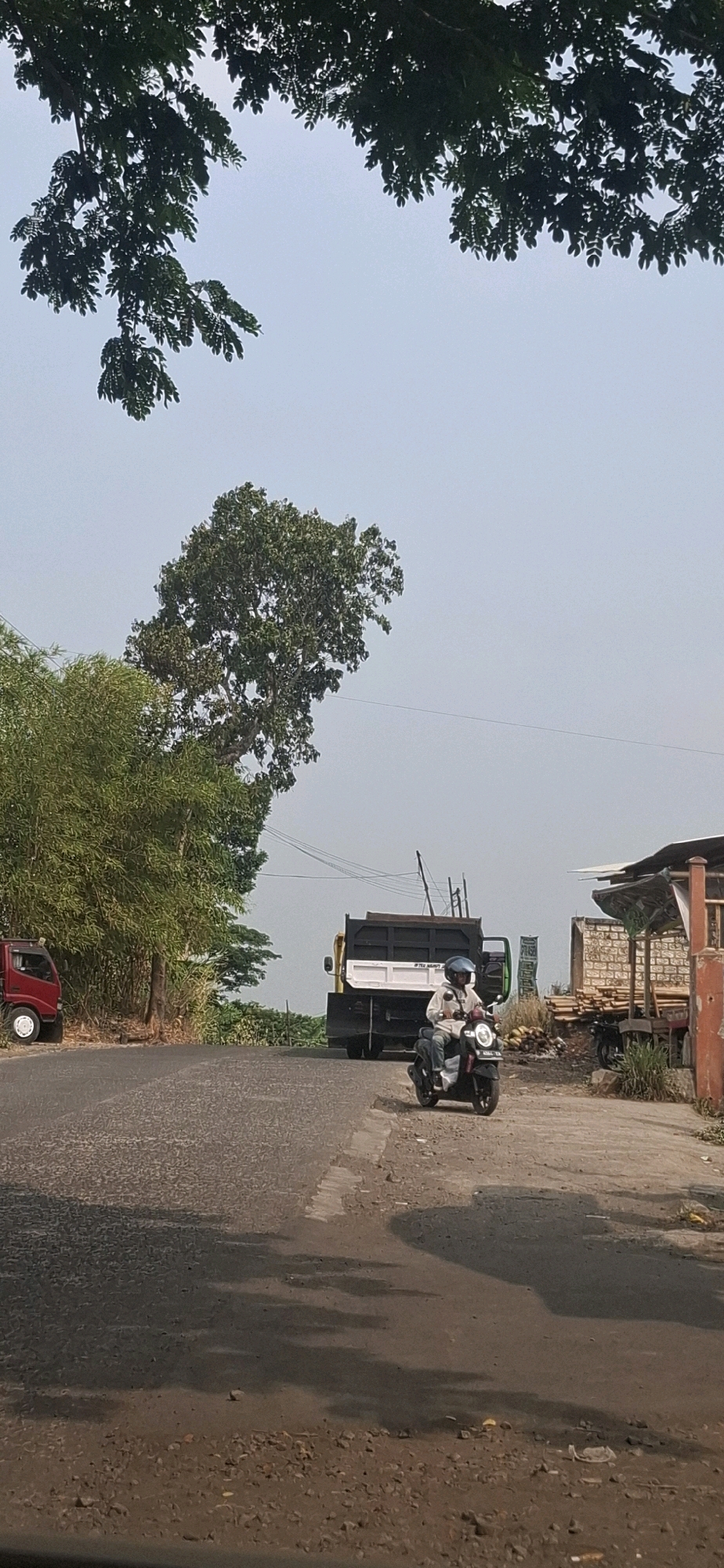 The image captures a scene on a paved road in Cibinong Hilir, Indonesia. On the left side of the road, a small red truck is parked. Further down the road, a large yellow and black dump truck is visible. In the foreground, a person is riding a dark-colored motorcycle, wearing a light-colored shirt, blue helmet, and jeans. The motorcycle is moving away from the viewer. The road surface appears weathered and cracked in some areas, with shadows cast across it. To the right of the road, there is a building under construction or with a rustic appearance, with piles of wood or building materials stacked nearby. Lush green trees and foliage frame the upper part of the image and line the left side of the road, suggesting a semi-rural or suburban environment. The sky is a pale, hazy blue, indicating a bright but potentially humid day.