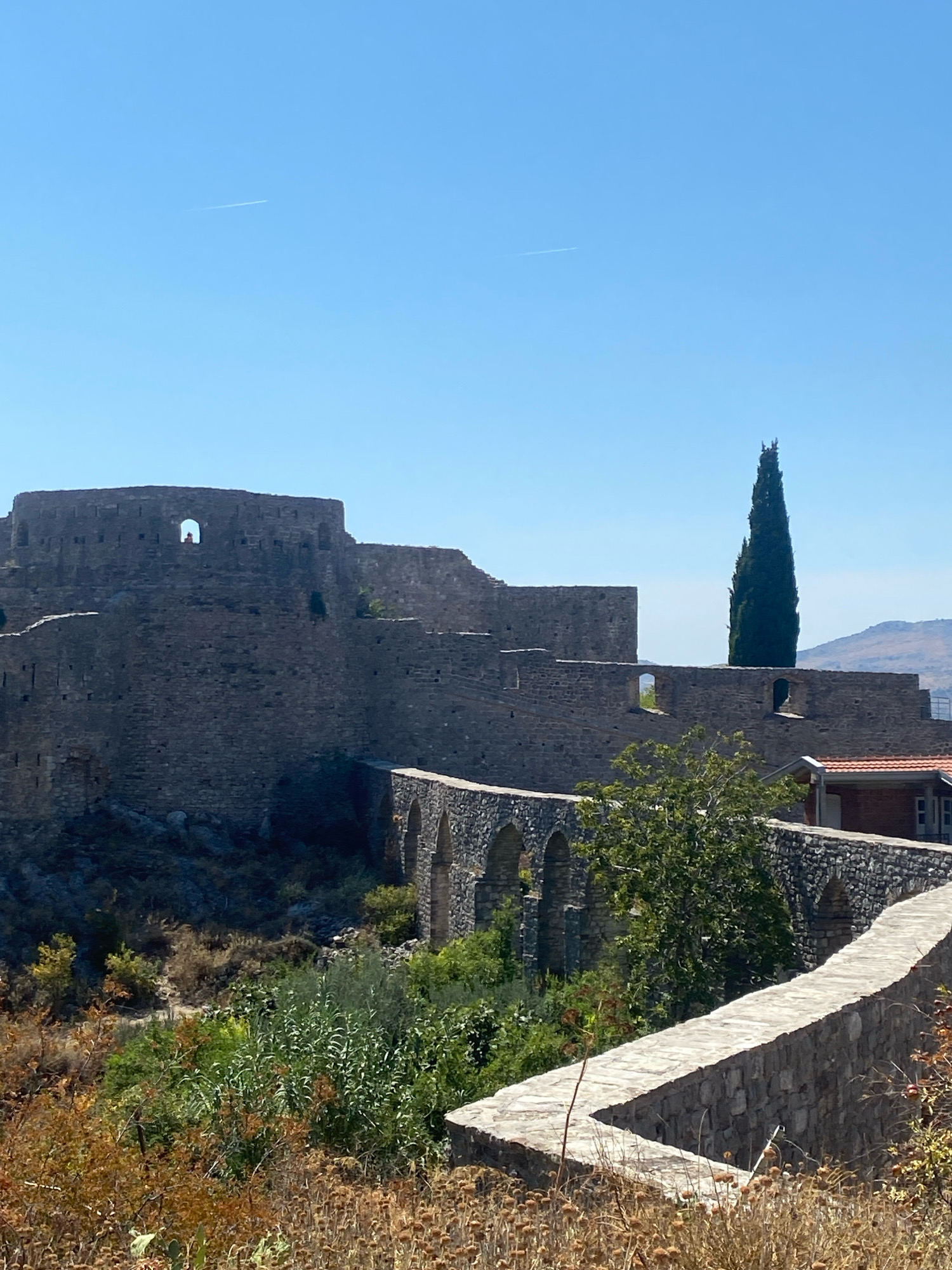 The image shows a view of the Stari Bar fortress in Montenegro. The fortress is made of stone and features a high wall with battlements and arched windows. A tall, slender cypress tree stands in the background, contrasting with the ancient stone structure. In the foreground, dry grasses and some green foliage are visible. A person is seen standing in an arched opening in the fortress wall. The sky is clear and blue, with two faint contrails from airplanes visible. The overall scene depicts a historical landmark under a bright, sunny sky.