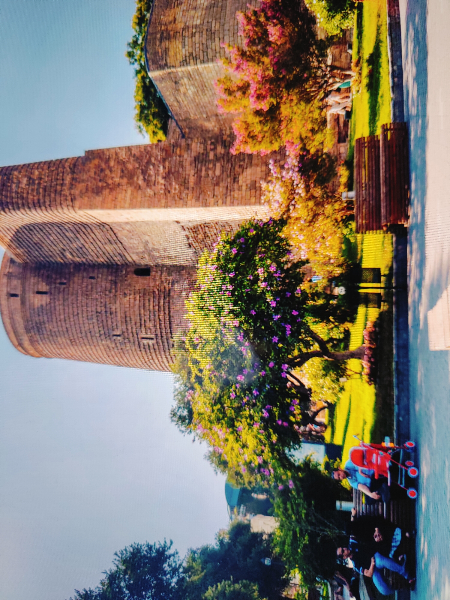 The image depicts a scene in Baku, Azerbaijan, featuring the Maiden Tower, a historic landmark. In the foreground, a young child is seated in a red stroller next to a bench where two adults are sitting. The stroller is positioned to the right of the bench. The Maiden Tower, constructed of reddish-brown bricks with visible horizontal lines, dominates the left side of the frame. Surrounding the tower is a park area with lush green grass, trees with vibrant pink and yellow foliage, and a clear blue sky. The park appears to be a public space with wooden benches and a paved walkway. The overall atmosphere suggests a pleasant day out in the city.