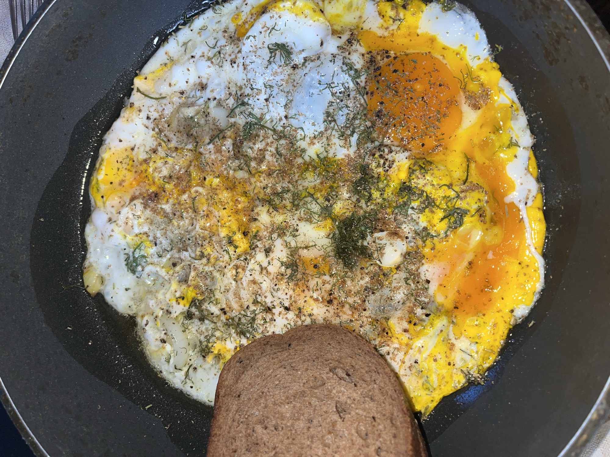 In Vasilyevo, Russia, a close-up, top-down view captures a frying pan on a stovetop. Inside the pan, a fried egg is being cooked. The egg has a partially set white and two distinct, bright orange yolks. Dried herbs and spices, likely dill and pepper, are sprinkled over the egg. To the left of the egg, a slice of dark rye bread sits in the pan. The pan itself is dark and appears to be non-stick, with a small amount of oil visible. The background is dark and out of focus, suggesting an indoor setting.
