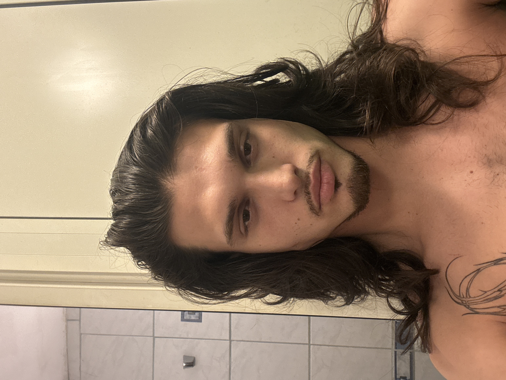 A young man with long, dark hair and a goatee is posing for a selfie in what appears to be a bathroom. He is looking directly at the camera with a neutral expression. His hair is slightly messy, with some strands falling over his face. He is shirtless, and a tattoo is visible on his chest. The background shows a tiled wall on the left, likely a shower or a bathroom wall, and a plain, light-colored wall on the right, possibly a door or cabinet. The overall lighting is soft and diffused. There is no indication of the city of Sofia, Bulgaria, in the image.