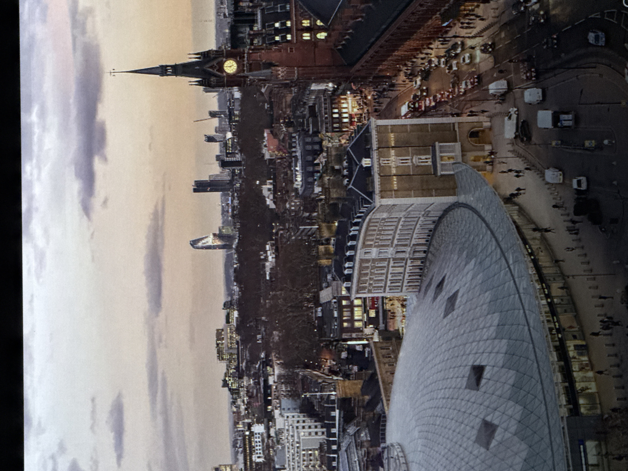 The image is an aerial view of a city skyline at dusk. The foreground features a large, domed glass structure with a patterned roof. Adjacent to this structure is a tall, dark-colored building with a clock tower, characteristic of Victorian architecture, which displays a illuminated clock face. To the right of the clock tower, a red brick building with many windows is visible. The middle ground shows a street lined with vehicles, including cars, vans, and what appears to be a red double-decker bus. Pedestrians are visible on the sidewalks and crossing the street. Buildings of varying heights and architectural styles populate the background, with some illuminated windows suggesting interior lighting. The sky is a muted orange and purple, with scattered clouds, indicating either sunrise or sunset.
The prompt specifically requests the location in Saratov, Russia, but the visual elements of the image, such as the clock tower resembling St Pancras clock tower in London, the red double-decker bus, and the overall architectural style, are not consistent with typical scenes in Saratov, Russia. Therefore, the image is likely not of Saratov, Russia.