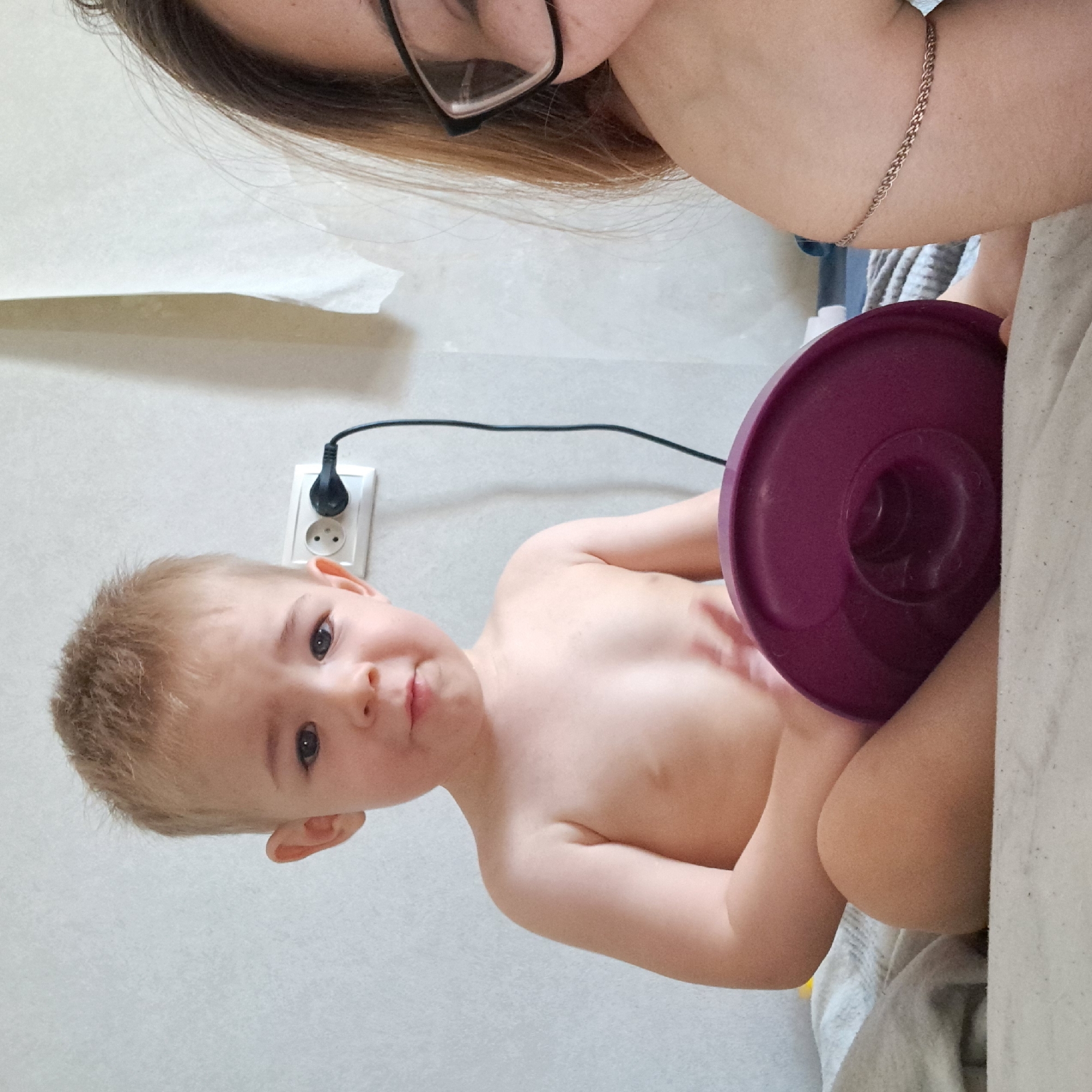 The image shows a young child sitting on a bed, holding a purple object. To the right of the child, a woman is partially visible, wearing glasses. An electrical outlet with a plug and cord is on the wall behind the child. The setting appears to be indoors, likely a bedroom. There is no information in the image that would indicate the location is the city of Krasnoyarsk, Russia.