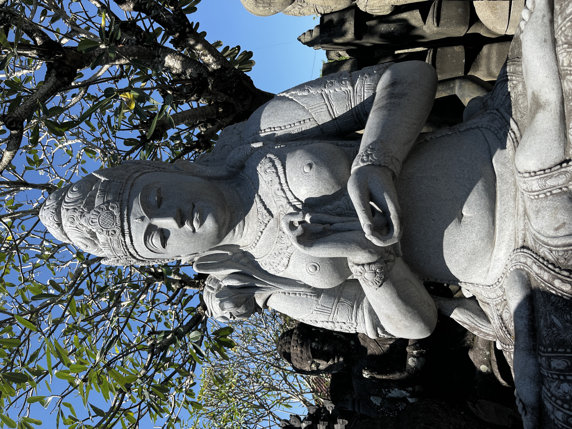 The image shows a stone statue of a seated female figure with hands pressed together in front of her chest, set against a bright blue sky and lush green foliage. The statue is intricately carved, with a detailed headdress, ornate jewelry, and flowing robes. Behind the main statue, other stone sculptures are visible, including a darker, possibly older, statue with a serene expression. The scene appears to be an outdoor setting, possibly a temple or a garden in Sukawati, Indonesia, a region known for its artistic traditions and stone carving. The sunlight casts shadows, highlighting the texture and form of the stone.