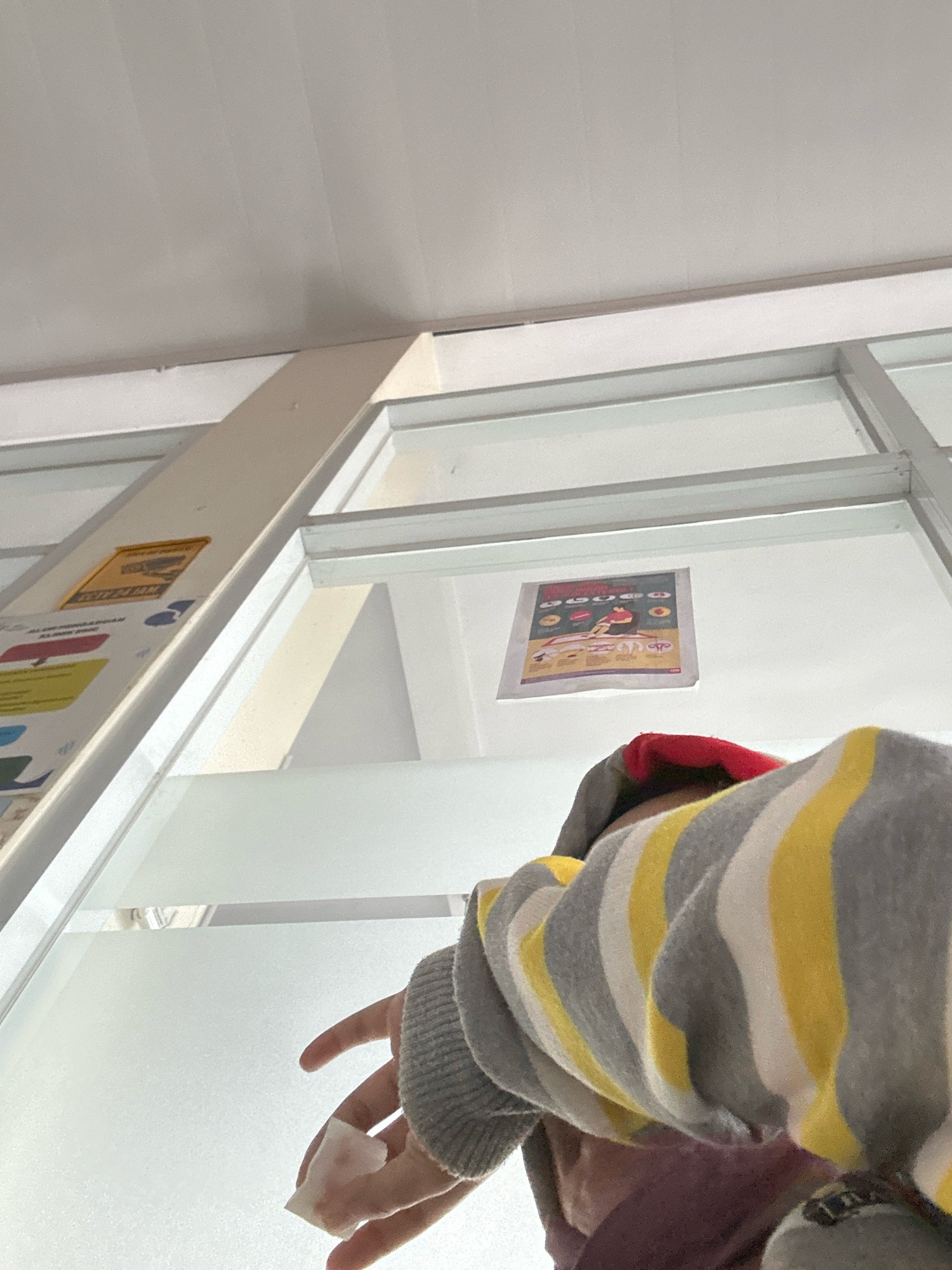 A low-angle, indoor shot captures a person's arm and shoulder in the foreground, wearing a gray, yellow, and white striped hoodie with a red lining visible around the hood. The person's hand, holding a white tissue, extends towards the left. The background features a frosted glass wall with a metal frame, extending upwards to a white ceiling with visible fluorescent light fixtures. To the left, a yellow sign with some text is attached to a vertical beam. Further back, on the glass wall, a colorful poster with images and text is displayed. The overall environment appears to be a public indoor space, potentially a clinic or educational institution, given the signage. The lighting is bright and diffused.