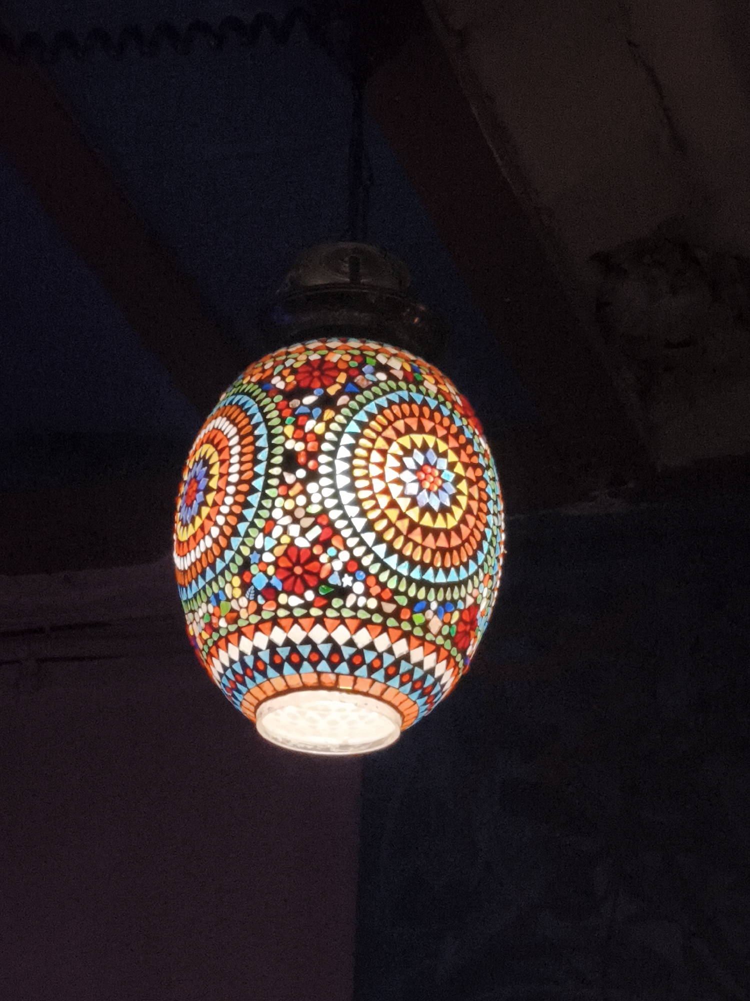 A dimly lit, medium-shot captures a vibrant, egg-shaped mosaic lamp hanging from a dark ceiling. The lamp features a complex pattern of multicolored glass pieces arranged in concentric circles and floral motifs. Colors include red, orange, yellow, blue, and green. The bottom of the lamp emits a bright, white light. The ceiling above is dark and includes wooden beams. The exact location in the city of Barcelona, Spain is not evident.