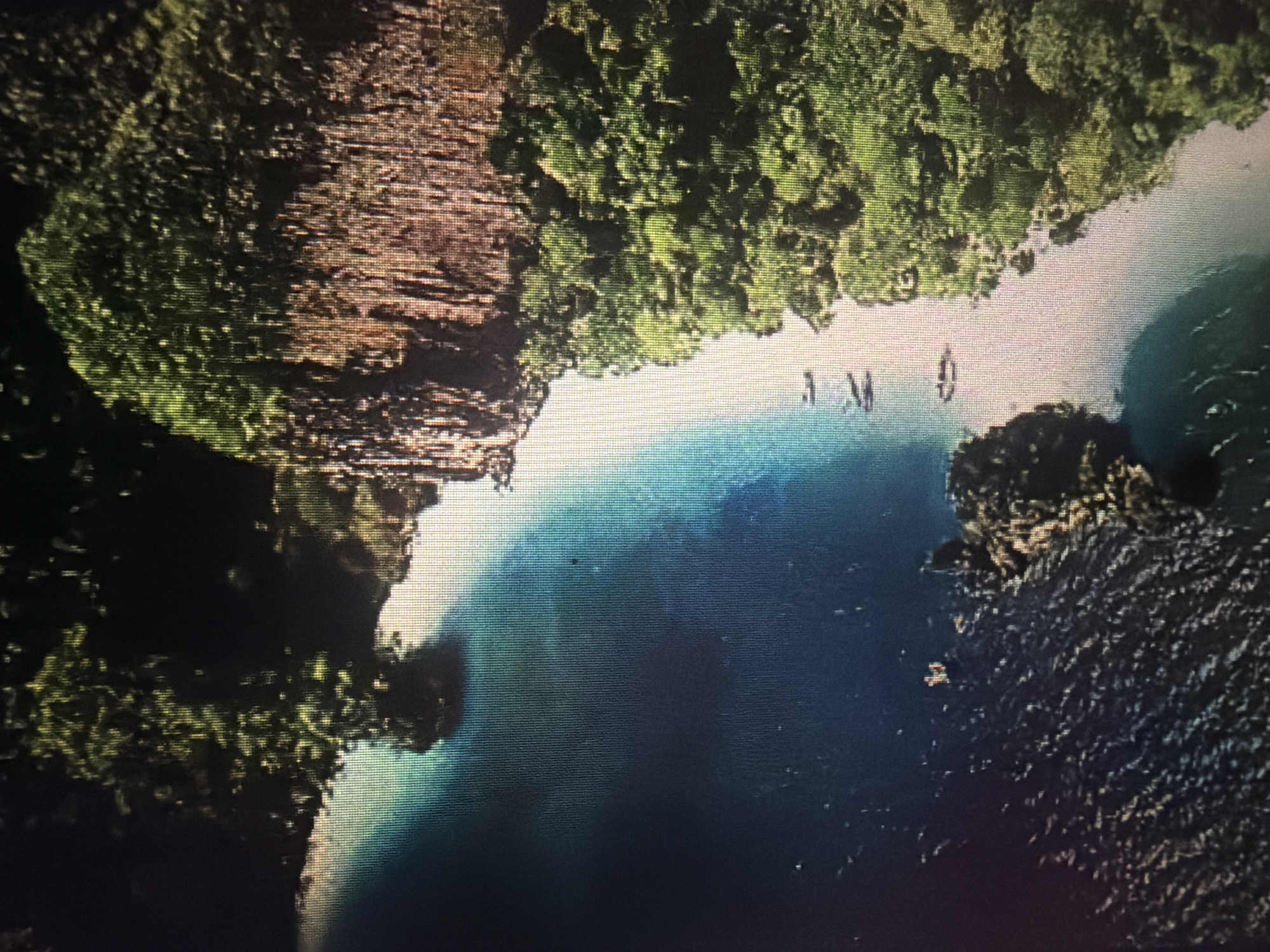 This is an aerial view of a tropical coastline. The scene features lush green vegetation covering steep cliffs and islands. The water is a vibrant turquoise near the shore, gradually deepening to a dark blue further out. Several small boats are visible on the water, some close to shore and others further out. One boat appears to be anchored near a rocky outcrop. The coastline is characterized by a white sandy beach separating the jungle-covered land from the sea. The overall impression is one of a serene and remote tropical paradise. The location is identified as Baan Mae Nam, Thailand, a coastal area known for its natural beauty.