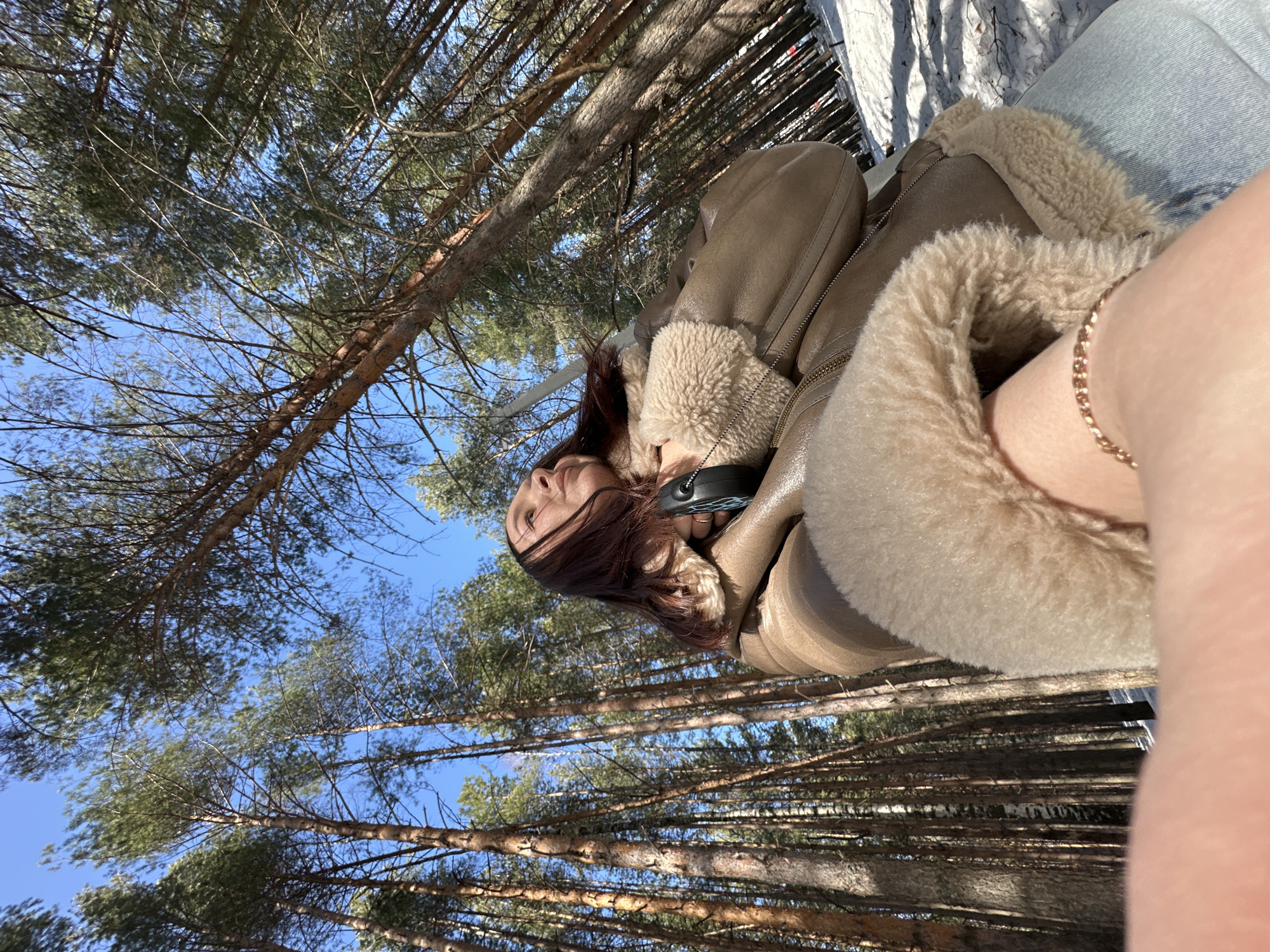 The image depicts a low-angle selfie portrait of a single female subject in a snowy forest within the city of Chaikovsky, Russia. The subject is positioned in the lower-left to mid-left frame, shown in profile with her head tilted back, gazing upwards and to the right. She has long, dark reddish-brown hair. She is wearing a light brown, possibly faux-shearling, jacket featuring prominent cream-colored fleece lining visible at the collar and cuffs, and light blue jeans. A black object, suspended from a strap, rests against her chest. Her left forearm, extended towards the viewer and adorned with a thin, gold-colored chain bracelet, is visible in the extreme foreground, indicating the selfie perspective. The background consists of a dense coniferous forest, primarily composed of tall, slender pine trees with reddish-brown trunks and green needles, extending upwards towards a clear, bright blue sky. The ground beneath the trees is covered in a layer of white snow, with shadows cast by the trees. The scene suggests a clear, sunny day in a winter or early spring environment.