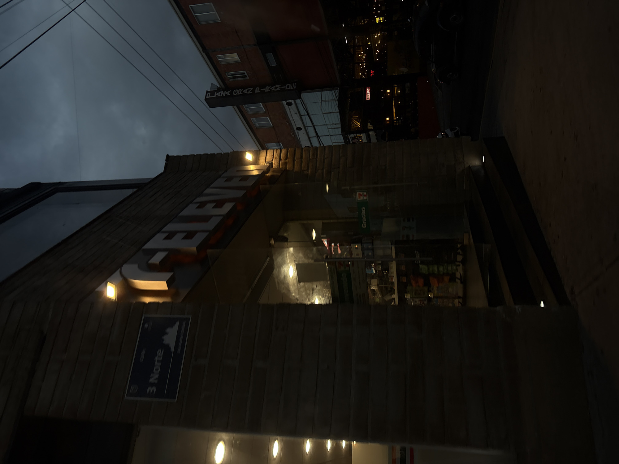 The image depicts an urban street scene at dusk or night in San Andrés Cholula, Mexico, specifically located at "3 Norte," as indicated by a blue street sign mounted on a building. The primary subject is the entrance of a 7-Eleven convenience store, constructed with a light-toned brick facade and illuminated by multiple warm-toned exterior spotlights. The store's interior is brightly lit, showcasing shelves stocked with products visible through its glass doors and windows. A green "Gracias" sign is affixed to the exterior glass, and the word "Bienvenido" is faintly visible inside. A series of dark steps leads from the concrete sidewalk up to the store's entrance.
Overhead, multiple power lines are visible against a dark, heavily clouded sky. To the right of the 7-Eleven, a multi-story building of red brick construction with several rectangular windows is present, featuring a tall, vertical sign that reads "PLAZA GRAN PIRAMIDE." Further down the street, additional commercial establishments are discernible, some adorned with decorative string lights; a "Tecate Box" sign is visible among them. A dark-colored vehicle, consistent with a Volkswagen Beetle, is positioned on the asphalt street. No individuals are observed in the scene.