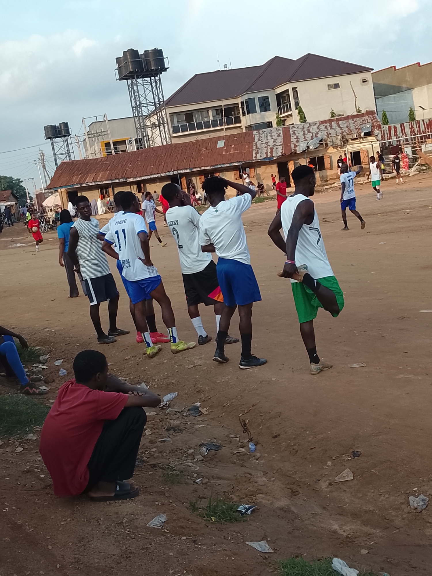 The image captures a group of young men engaged in a football (soccer) activity on a dusty, unpaved surface. The setting appears to be an urban area, likely a neighborhood in Abuja, Nigeria, characterized by informal structures and a somewhat unkempt environment. In the background, there are buildings, some with corrugated iron roofs, and prominent water towers. The sky is overcast, suggesting it might be late afternoon or early evening.
Several individuals are visible, most of whom are wearing sportswear, including jerseys and shorts, indicative of a sports practice or game. One person in the foreground, sitting down and facing away from the camera, is wearing a red t-shirt and dark pants. Other individuals are standing and interacting, with some seemingly involved in stretching or preparing for activity. One person is in the process of stretching their hamstring. The ground is sandy and uneven, with scattered debris, including plastic bags and wrappers, common in such environments. The overall atmosphere suggests a casual, community-based sporting event taking place in a typical Nigerian urban setting.