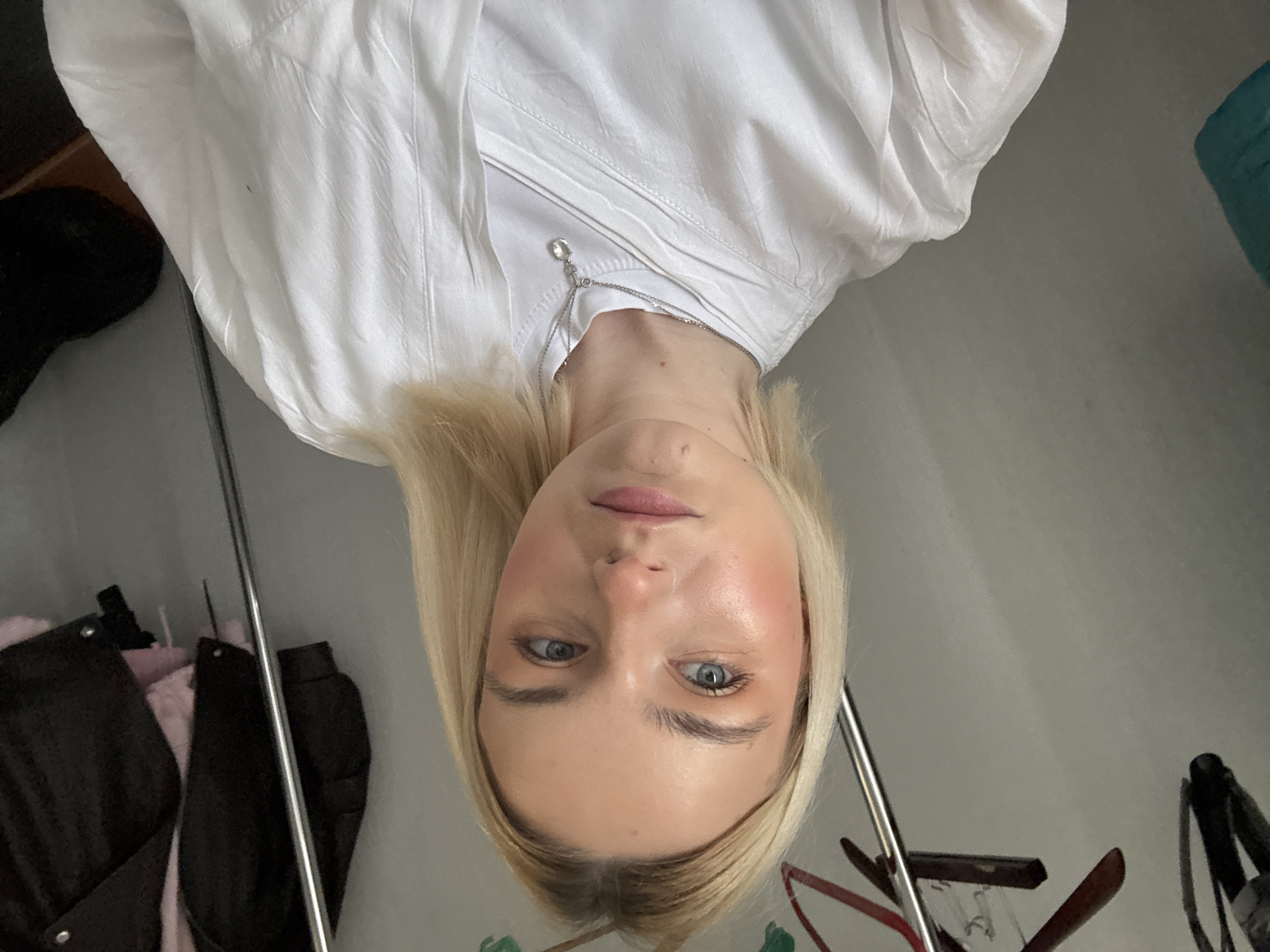 A young, fair-skinned woman with blonde hair and blue eyes is the focal point of this indoor photograph. She is wearing a white jacket over a white shirt, adorned with a delicate silver necklace.
In the background, clothing hangs on a rack, including a black coat and a pink garment. The location is uncertain but may be Saint Petersburg, Russia, given the possible reference in the prompt. The image appears to be taken from a slightly angled, tilted perspective, suggesting a selfie or a candid shot.