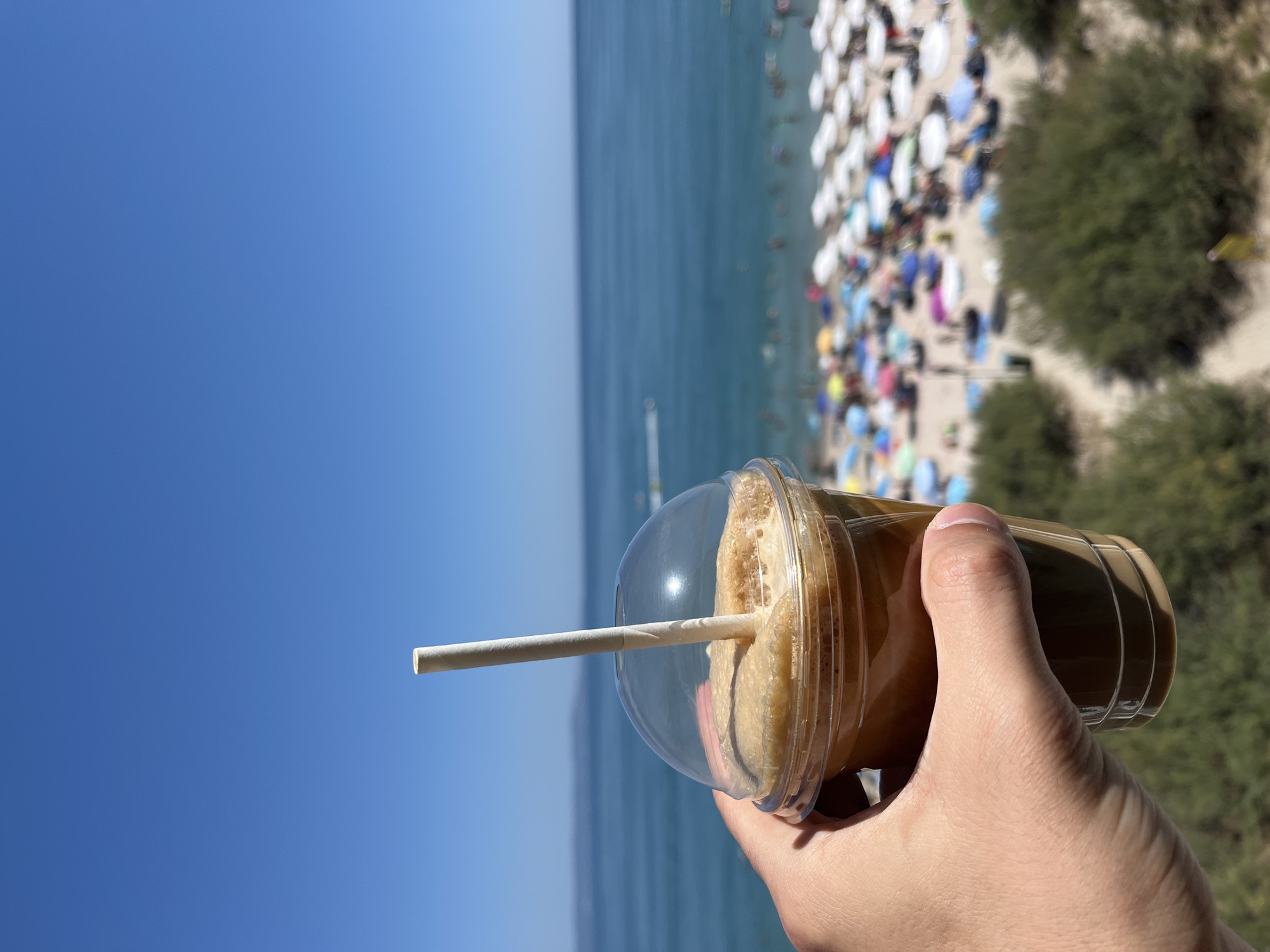 A close-up shot focuses on a hand holding a clear plastic cup filled with a frothy, light brown beverage and a light-colored paper straw. The background, out of focus, reveals a bustling beach scene in Pomorie, Bulgaria. Numerous beach umbrellas, predominantly white, with some colorful ones interspersed, dot a sandy beach. People are visible, some sitting or standing near the umbrellas, while others are in the turquoise sea. A thin strip of distant land or a peninsula is visible on the horizon under a clear, bright blue sky. Green shrubs are present in the foreground, suggesting a slightly elevated vantage point overlooking the beach.