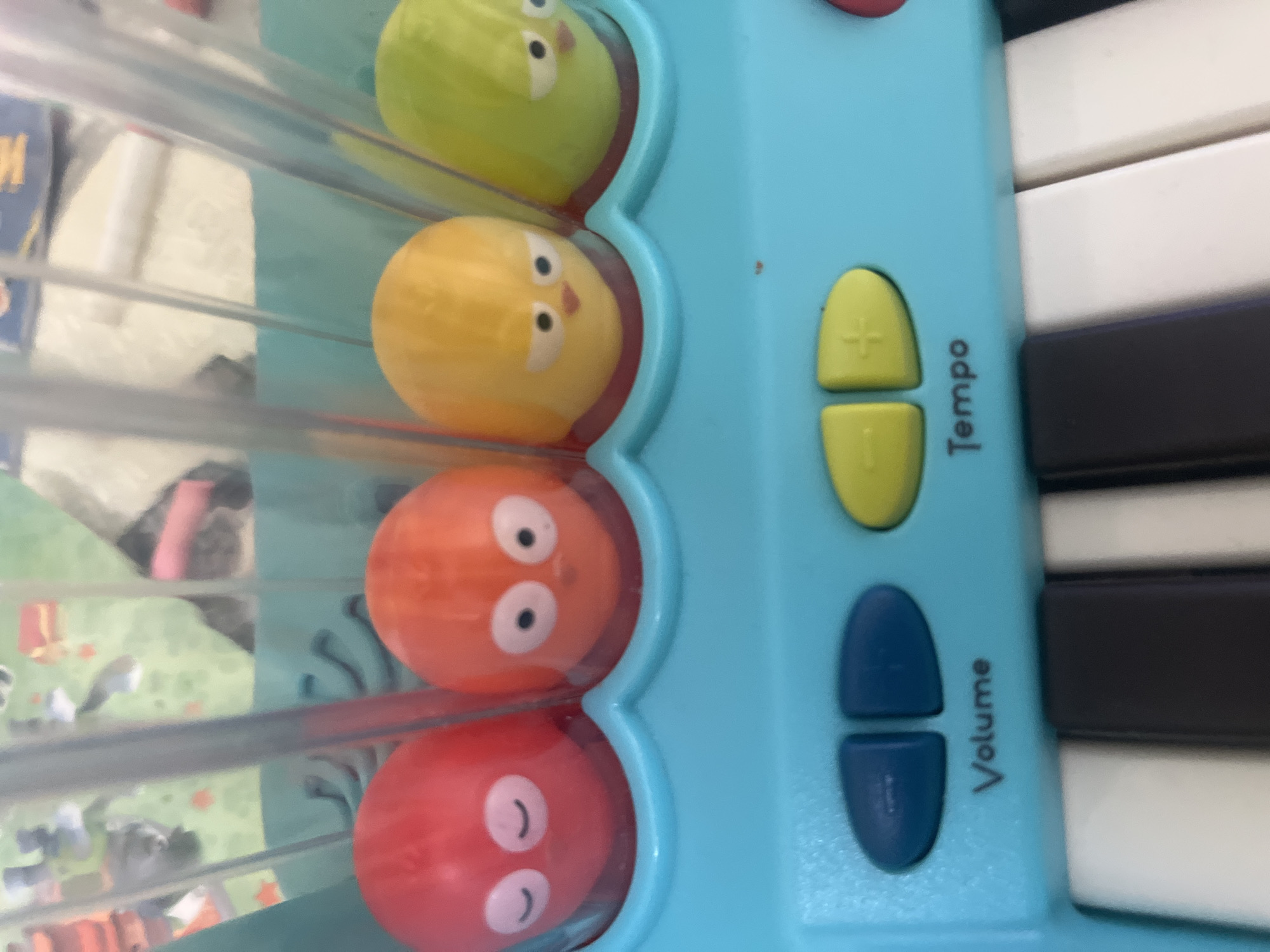 The image displays a close-up, rotated view of a colorful children's musical toy, likely a toy piano or accordion. The main body of the toy is made of light blue plastic. On the left side, four spherical, cartoon-like characters are arranged vertically within clear plastic enclosures. From top to bottom, these characters are green, yellow, orange, and red. The green and yellow characters have small black dot eyes and beaks, while the orange and red characters feature larger white eyes with black pupils and subtle mouth shapes.
To the right of these characters, on the blue plastic surface, are control buttons. Two yellow oval buttons, marked with a plus (+) and minus (-) sign, are accompanied by the vertical text "Tempo." Below these are two blue oval buttons, next to which the word "Volume" is printed vertically. To the far right, sections of white and black piano keys are visible, indicating the toy's function.
The background, seen through a clear plastic panel, is out of focus but suggests an indoor setting, possibly a child's room or play area. Blurry shapes in the background include what appears to be fabric or a patterned surface with cartoon figures. There are no people visible in the image. The only visible text on the toy itself reads "Tempo" and "Volume." The location context is Onokivtsi, Ukraine.