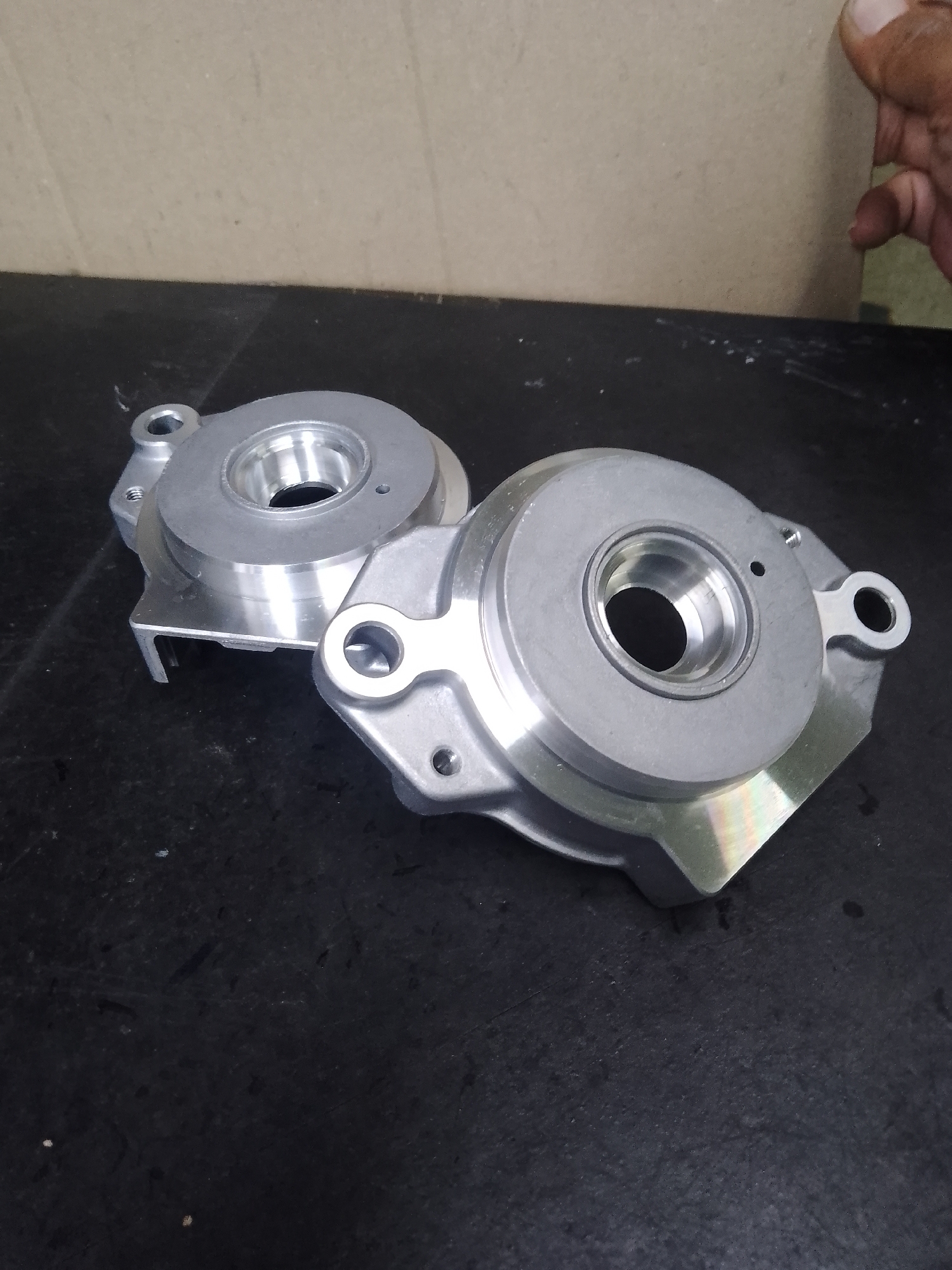 The image shows two metallic objects on a dark, textured surface, likely a table or workbench. The objects are identical, appearing to be some type of mechanical component or housing, possibly for machinery or automotive parts. They are made of a polished, silver-toned metal, with intricate machining evident. Each object features a circular opening in the center, with raised lips and internal detailing. There are also mounting holes and projecting flanges.
The background is a mottled, light brown surface, possibly a wall or partition, with a hint of a cardboard-like texture. In the upper right corner, a person's hand is partially visible, with fingers extended towards the background surface, suggesting someone is holding or interacting with it.
The lighting is somewhat diffused, creating highlights and shadows on the metallic objects, which emphasize their three-dimensional form and machined surfaces. There are some small white specks and smudges on the dark surface, indicating a workshop or industrial setting. The overall impression is of manufactured parts ready for assembly or inspection. No text is visible in the image. The scene appears to be indoors, in what could be an industrial or manufacturing environment in Ciantra, Indonesia.