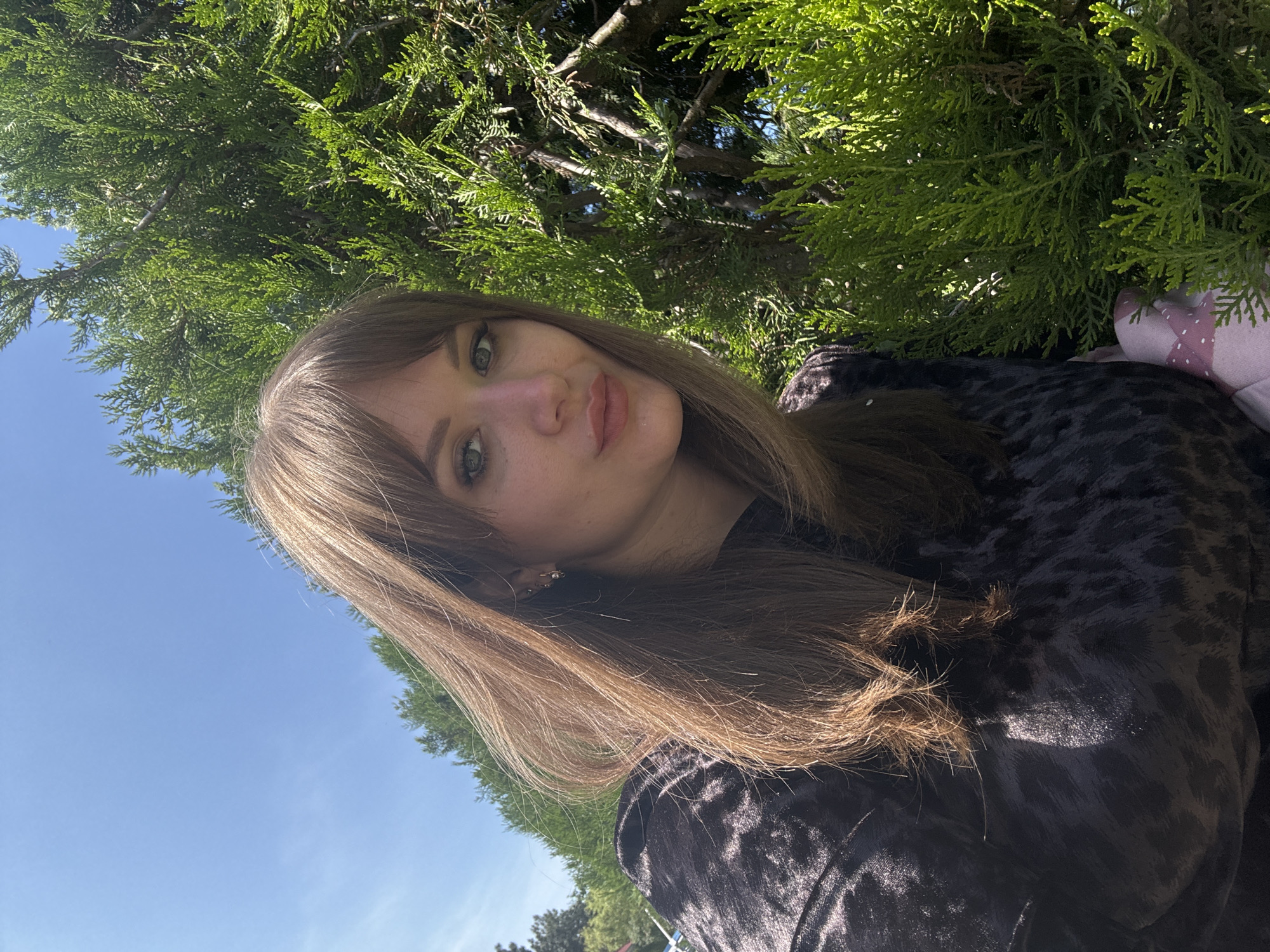 A young woman with long brown hair and green eyes is posing for a selfie outdoors. She is wearing a dark patterned shirt and standing in front of a lush green tree. The sky is visible in the background and is a clear, bright blue. A pink object with white dots is visible on the right. The location is not discernible from the image.