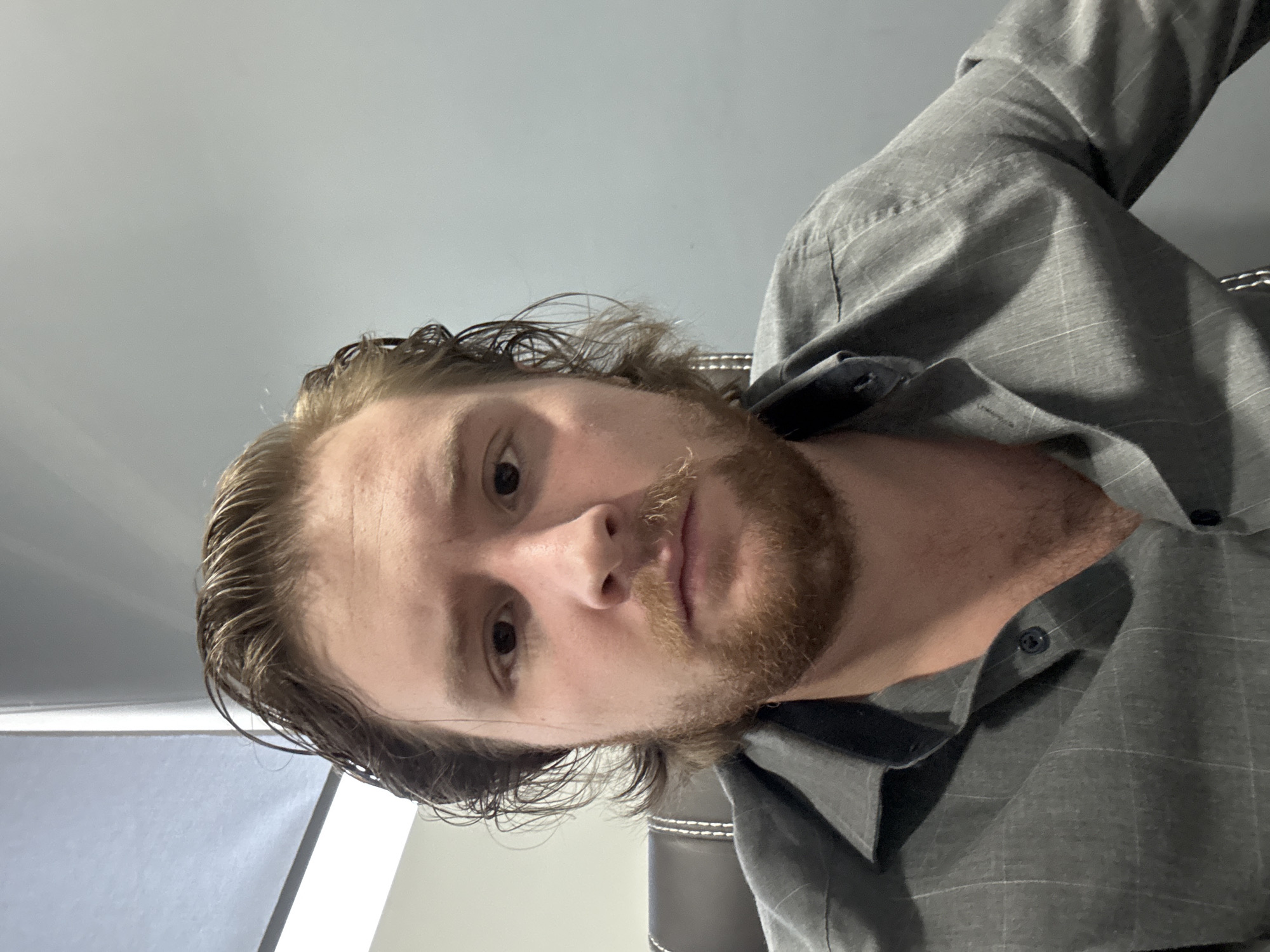 A fair-skinned man with light brown hair and a beard is shown from a low-angle perspective. His hair appears wet. He is wearing a gray, button-down shirt. The background consists of a plain gray wall and what appears to be a window or light source. The location is likely indoors, but there are no details to suggest it is in Cuautitlán Izcalli, Mexico.