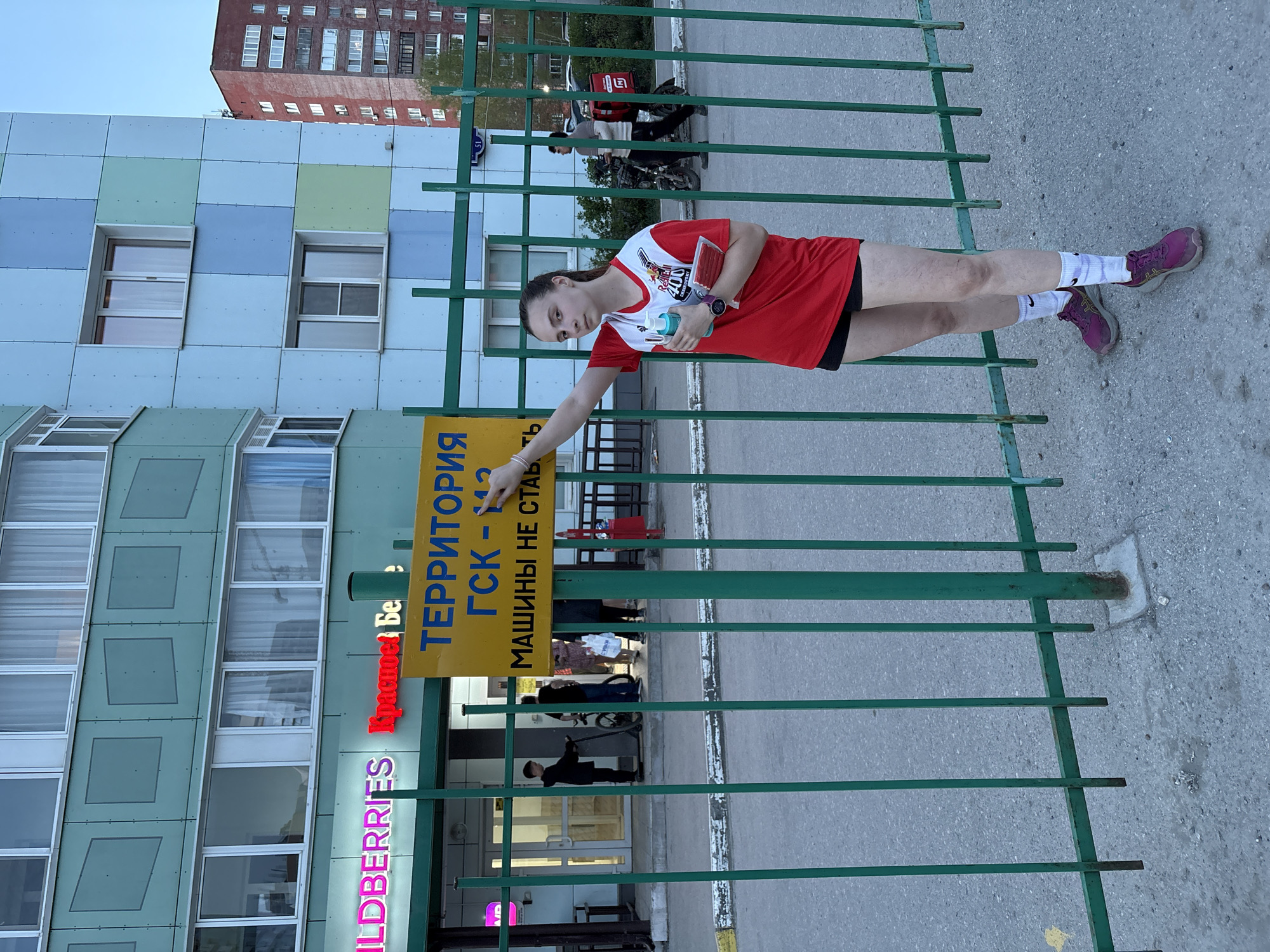 A young woman stands next to a green metal fence in Perm, Russia. She is wearing a red and white athletic shirt, black shorts, white socks, and purple running shoes. She holds a water bottle and what appears to be a packet in her hands and points to a yellow sign attached to the fence. The sign reads "ТЕРРИТОРИЯ ГСК-12 МАШИНЫ НЕ СТАВ". The sign is in Russian and translates to "Territory GSC-12 Cars Not Allowed." Behind the fence is a building with a "WILDBERRIES" store sign. People can be seen entering and exiting the store. In the background are apartment buildings. A delivery person on a scooter is visible further back. The lighting indicates it's daytime.