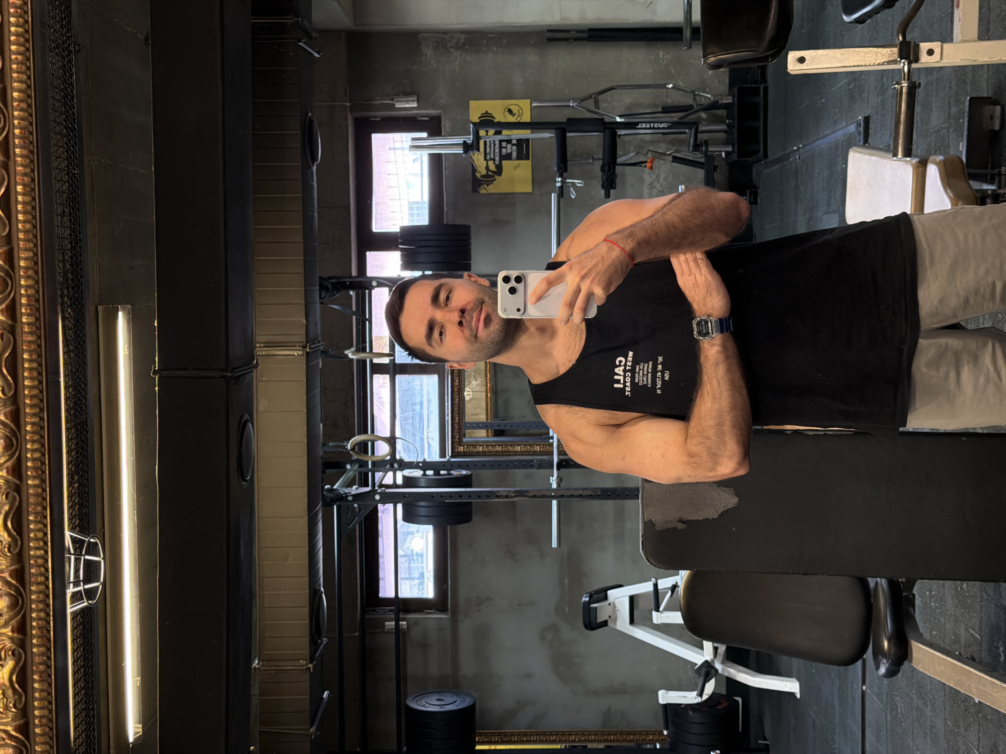 A man takes a selfie in a gym in Tbilisi, Georgia. The man is light-skinned with short dark hair, wearing a black tank top and grey shorts. He is holding a white smartphone, taking a selfie with his left hand. He is wearing a blue watch on his left wrist and a red string on his right wrist. The gym is located in an older building with exposed grey concrete walls. There are weightlifting machines and equipment throughout the gym. There is a squat rack with weight plates loaded on the bar. There are also several windows in the background, with light streaming in.