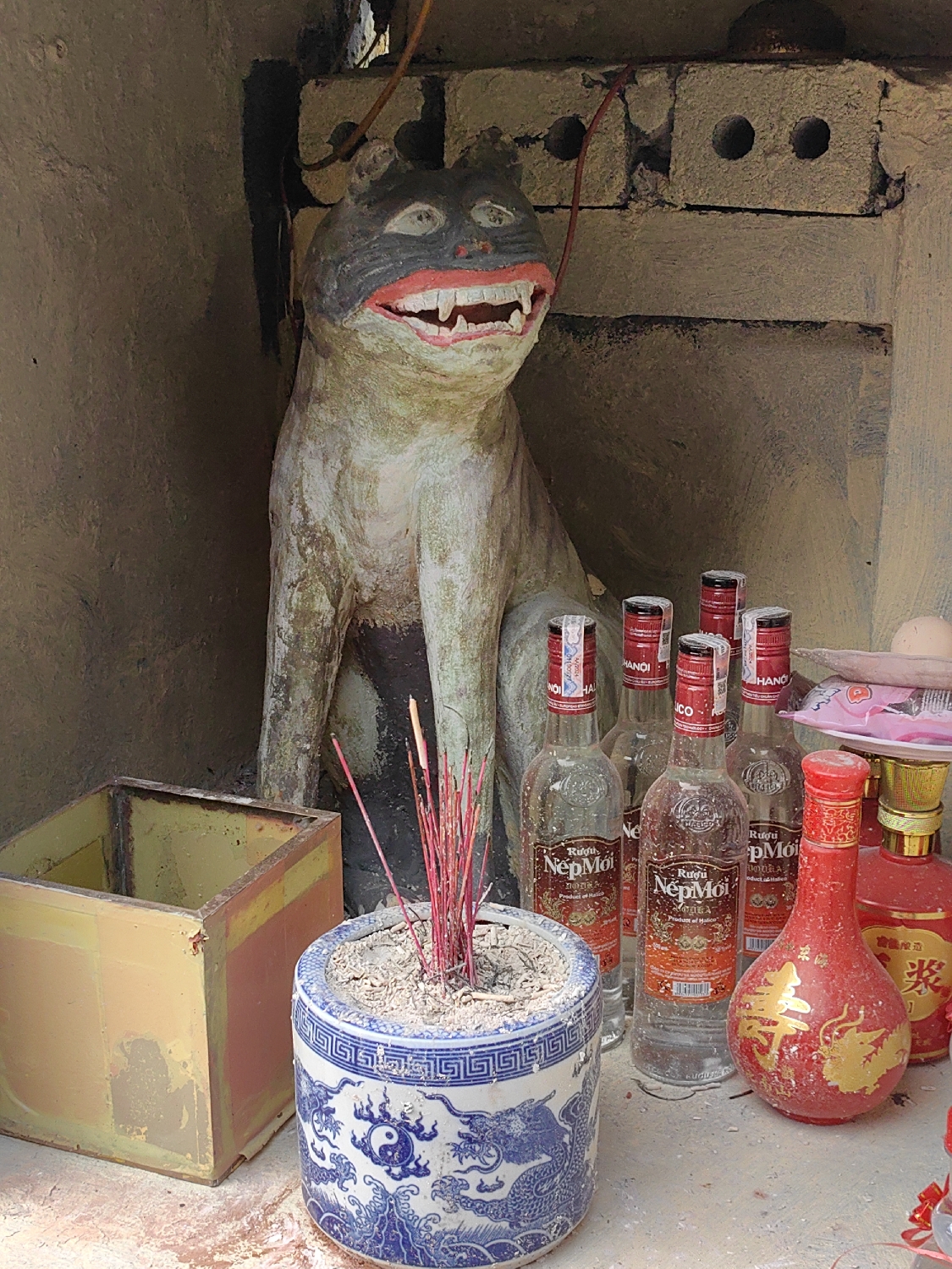 An indoor or shaded scene depicts a small, folk-art shrine, likely in Bai Chay Ward, Vietnam. The central figure is a weathered, light-colored statue resembling a grinning cat or canine, with a dark, possibly black face, prominent red lips, and large white teeth. It sits upright against a backdrop of rough, light-colored brick or stone walls, with some exposed wiring visible above.
In front of the statue, various offerings are arranged. A blue and white ceramic incense pot, decorated with dragons and a yin-yang symbol, holds numerous red incense sticks, some appearing burnt. To its right, several clear glass bottles of alcohol are lined up. Their labels clearly show "Rượu Nếp Mới," "Product of Vietnam," and "HANOI." Adjacent to these, two ornate red bottles, one with gold decorative characters, are also present. To the left of the incense pot, a rectangular, yellowed metal box sits on the ground. The arrangement suggests a small altar or shrine for traditional spiritual practices.