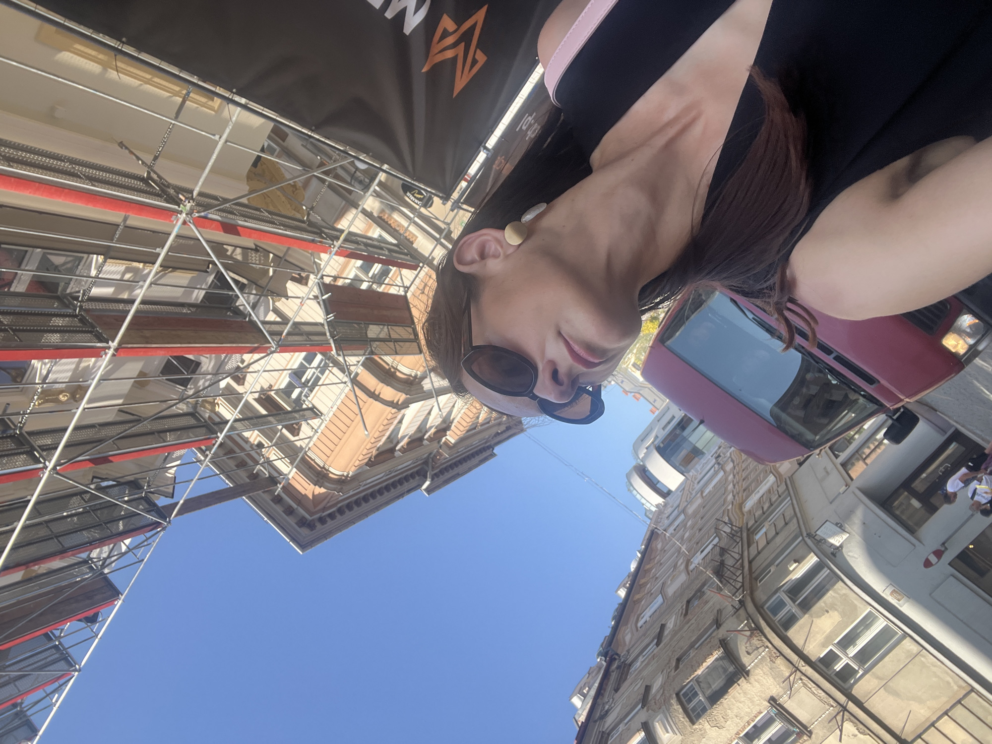 A low-angle, wide shot captures a woman in sunglasses and a black dress standing in front of an orange building covered in scaffolding under a clear blue sky. The woman is facing upwards, looking away from the camera, with her right shoulder and upper chest visible. Her brown hair is down, and she is wearing large, dark sunglasses and minimalist earrings. To her left, a red van is parked on a street, and in the background, a weathered, multi-story building is visible. A black banner with an orange geometric logo is partially visible in the lower right corner. The scene appears to be in an urban environment, possibly undergoing renovation.