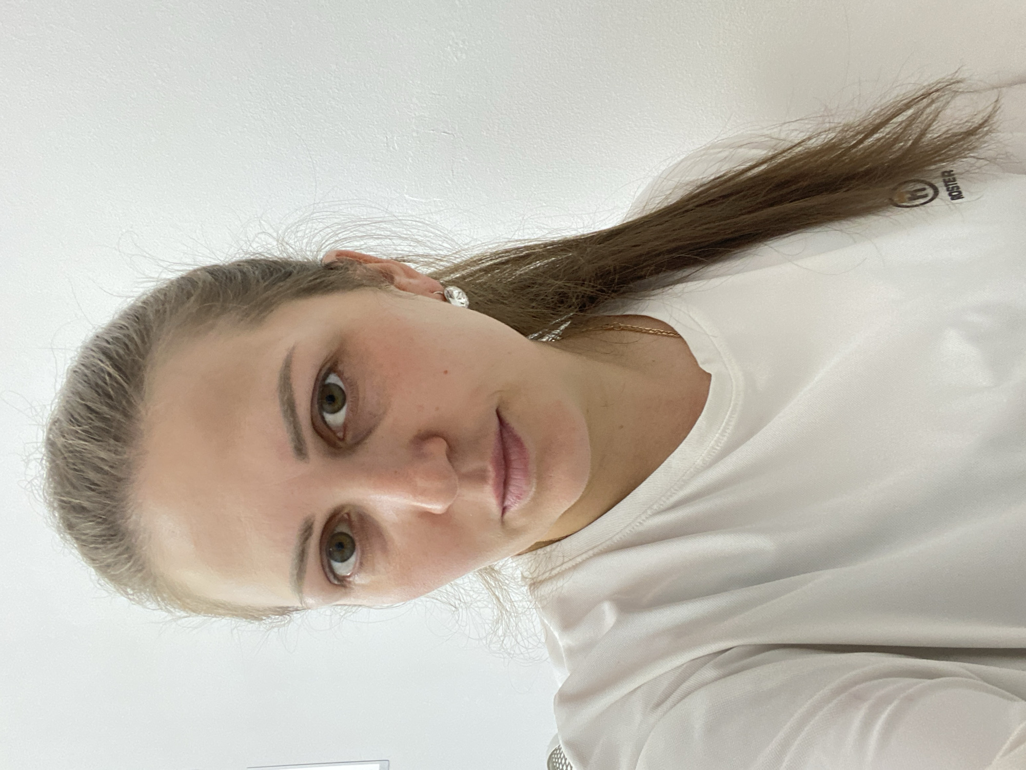 A woman is looking directly at the camera. She has fair skin, brown hair pulled back, and green eyes. She is wearing a white t-shirt and earrings. The background is a white wall. It is impossible to verify the location as Марковский, Russia based on the image.