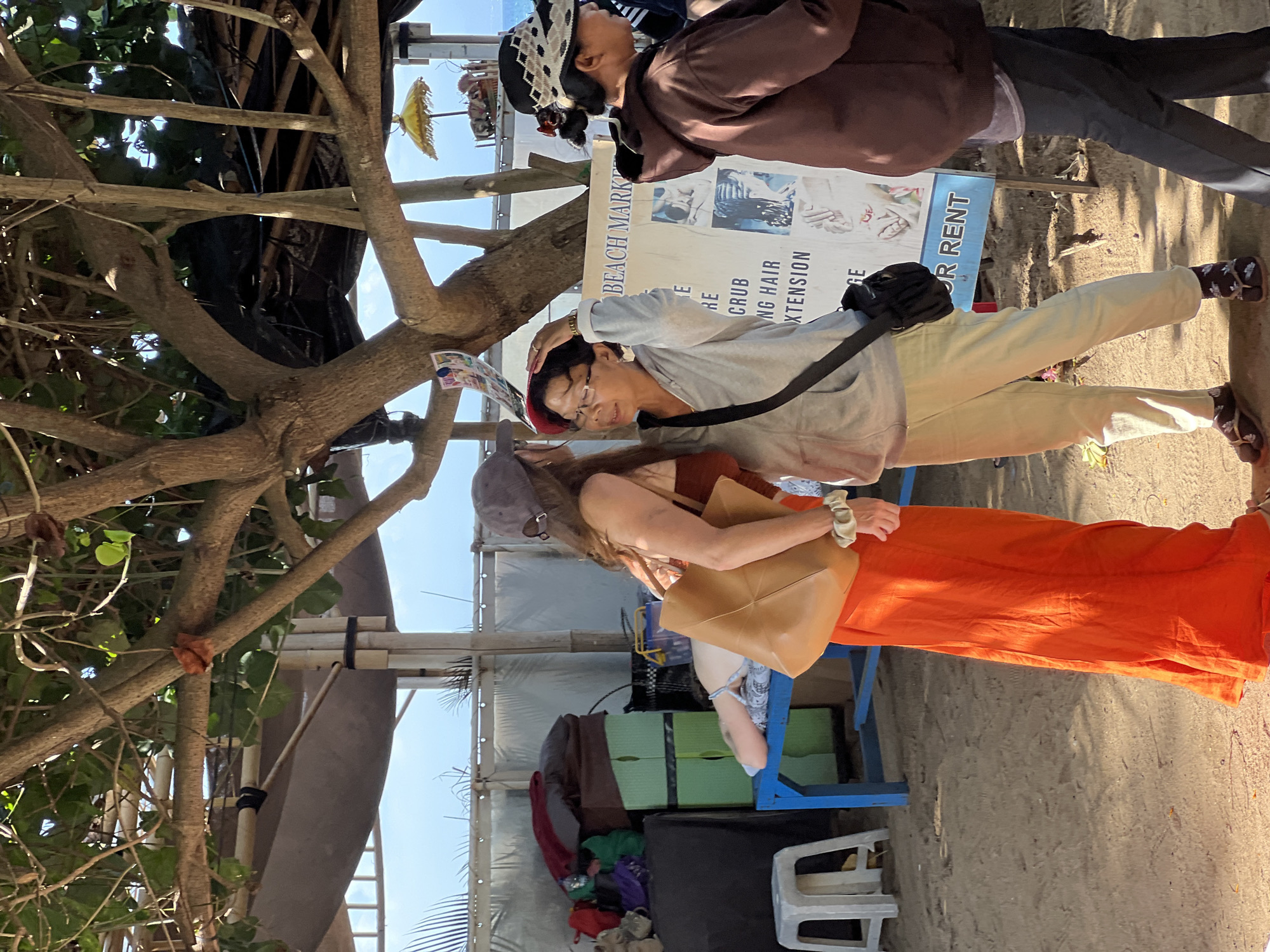 The media file depicts an outdoor scene in Denpasar, Indonesia. It features at least three individuals, two women and one person partially obscured. The person in the foreground, wearing glasses and a light-colored hoodie, is touching their head. They are dressed in light-colored pants and a black crossbody bag is visible. To their right, another woman, with a tan bag slung over her shoulder, is wearing a bright orange garment that appears to be a long skirt or sarong. Behind them, another person is dressed in a brown jacket and dark pants, also touching their head.
The setting appears to be a beach or a market area, with sand on the ground. A tree with thick branches partially obstructs the view, and in the background, there are structures that suggest stalls or temporary shelters, with some items like green plastic boxes stacked. There is a banner visible that appears to be advertising services. The text "BEACH MARK" and "FOR RENT" are partially legible on the banner, along with other words like "RE" and "GRUB" and "NG HAIR" and "XTENSION". A small yellow umbrella is visible further in the background. The lighting suggests it is daytime, with bright sunlight casting shadows. The overall atmosphere seems casual, possibly a busy market day.