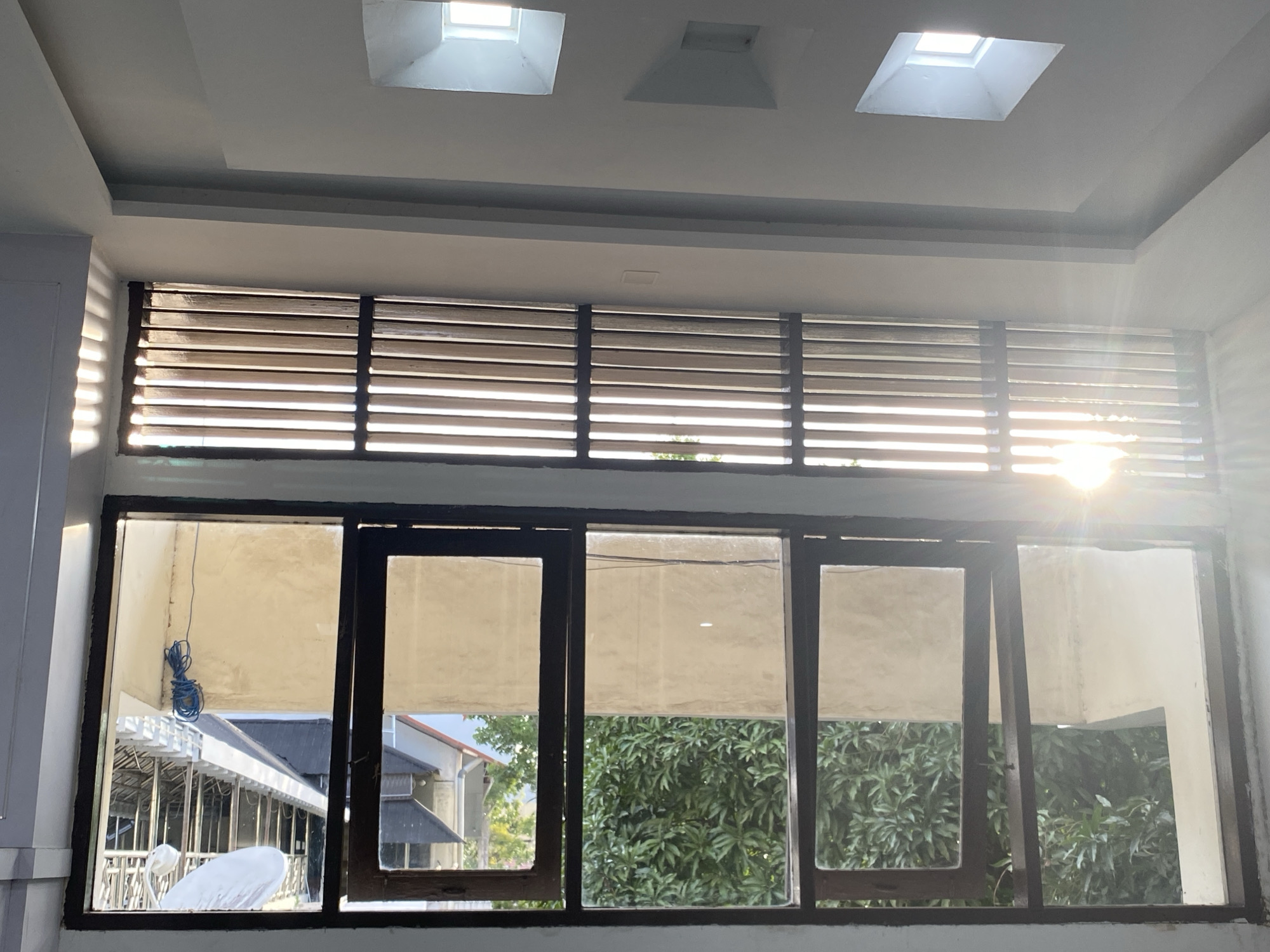 The image shows a view from inside a room looking out a large window. Above the window, the ceiling features a recessed design with three skylights. The window itself is divided into multiple panes by dark wooden frames. Horizontal blinds are visible across the top portion of the window, with sunlight streaming through them, creating distinct light patterns. Below the blinds, the lower portion of the window reveals an outdoor scene.
In the outdoor view, the foreground shows the interior of the room with beige-colored blinds or curtains partially drawn. A blue coiled wire is hanging on the left side. Beyond the window, there is an outdoor area with some structures, including a covered walkway with white pillars and a roof, and what appears to be a white, rounded object. Further in the background, there are glimpses of residential buildings with tiled roofs and dense green foliage from trees. The overall impression is of a residential or possibly commercial building with a view of a tropical environment. Given the prompt's request for the location of Banda Aceh, Indonesia, this scene is consistent with the architecture and vegetation found in that region, known for its warm climate and lush greenery. The image is captured during daylight, with the sun being a prominent light source.