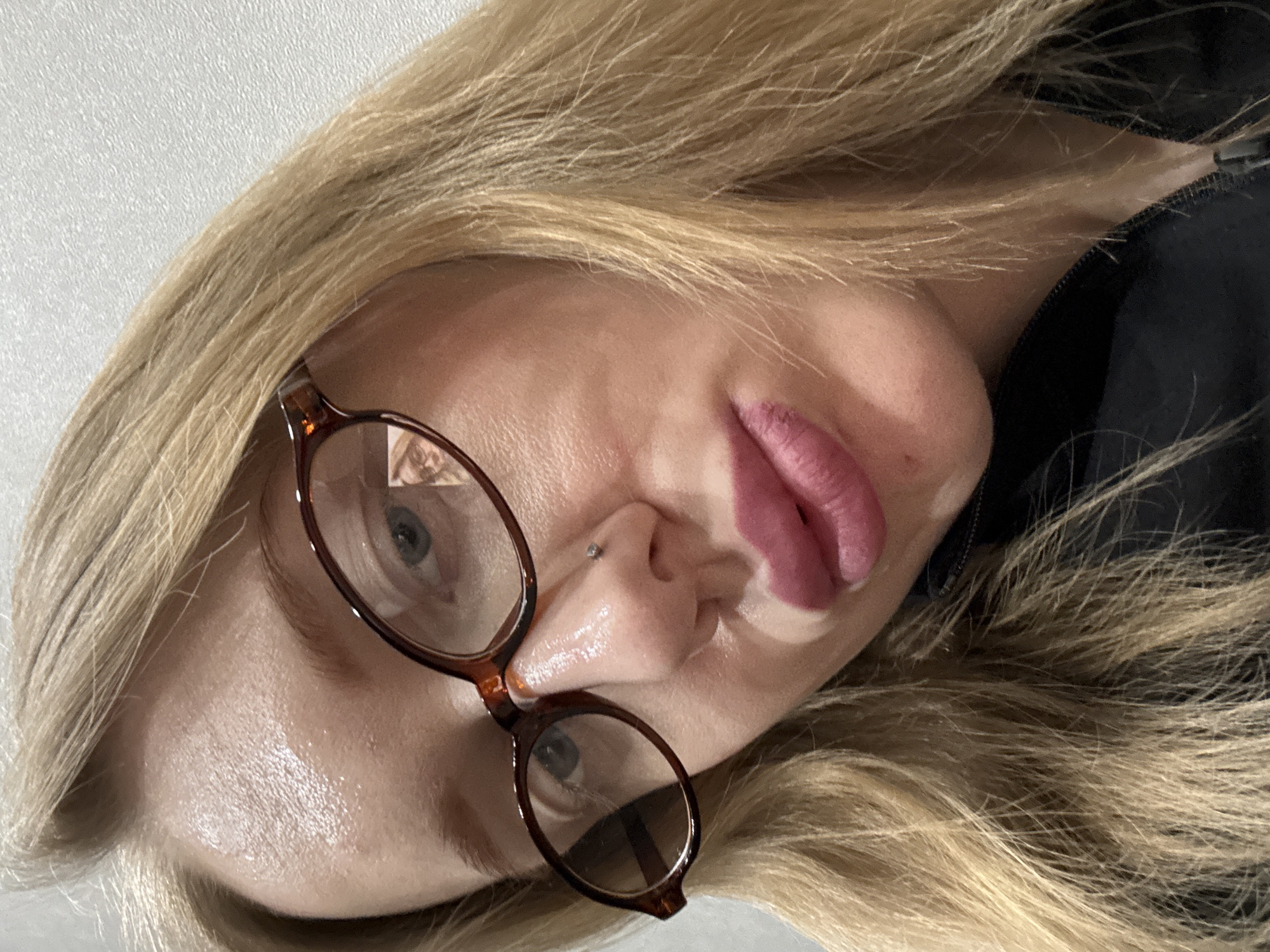 A close-up, slightly angled view shows a woman's face. She has blonde hair and is wearing glasses with brown frames. She has light skin and pink lips. A nose ring is visible on the left side of her nose. A portion of a black garment is visible on the right. The location in Kuleshovka, Russia is not discernible from the image.