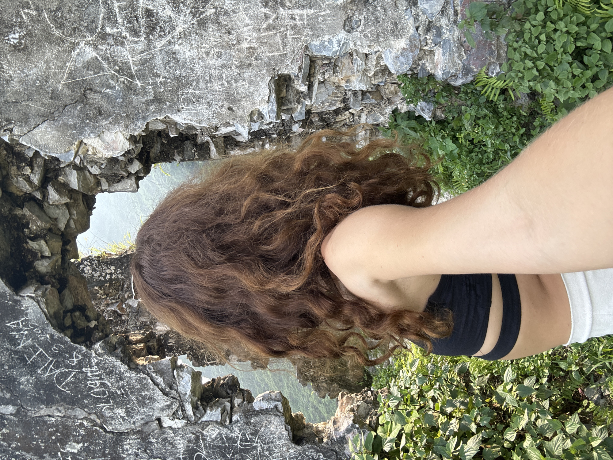 A woman with long, curly brown hair is positioned with her back to the viewer, standing atop a stone structure in Đường Thượng Commune, Vietnam. She wears a black top and white pants. The structure appears to be part of a ruined stone building or wall, as indicated by the rough-hewn stones and etched markings on the surface. Below, a dense layer of green vegetation fills the background and extends toward the edge of the frame. The perspective suggests the woman is standing at a considerable height, looking down upon the landscape.