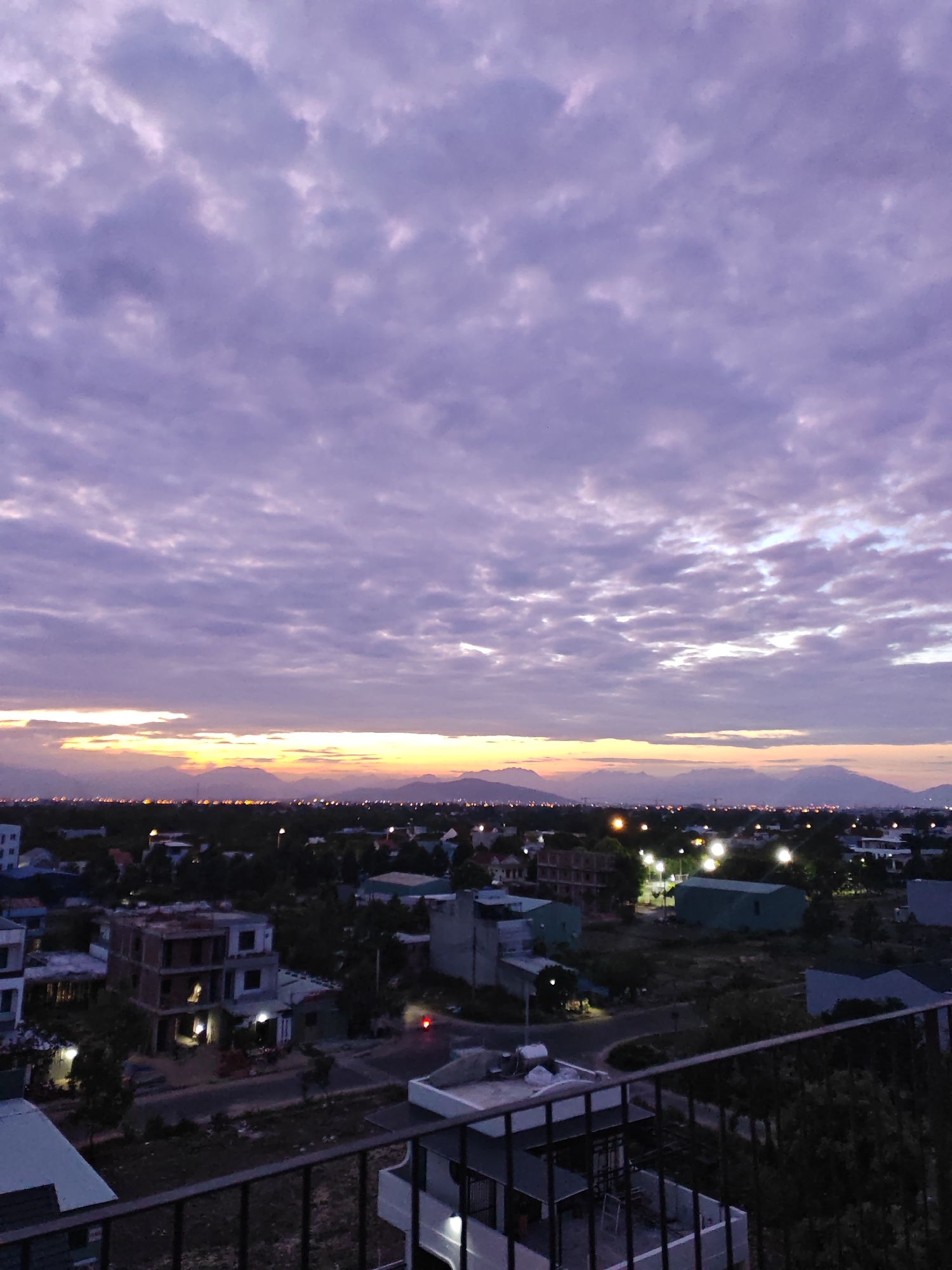 The image captures an expansive view of a city at dusk, likely from an elevated vantage point, overlooking rooftops and a distant mountain range. The sky is a prominent feature, filled with a textured canvas of purplish-gray clouds, suggesting either twilight or an overcast evening. A sliver of bright, yellowish light is visible on the horizon, bordering the dark silhouettes of the mountains, indicating the sun is either setting or has just risen. Below the sky, the urban landscape unfolds. Buildings of various sizes and architectural styles are scattered across the scene, some appearing unfinished with exposed concrete structures, while others are complete with dark roofs and visible windows. Streetlights and lights from within some buildings cast a warm, diffused glow, illuminating the city's contours. The overall impression is a quiet, developing urban environment under a dramatic sky. The presence of construction sites and what appear to be low-rise residential and commercial buildings suggests a developing area. Without specific landmarks, pinpointing the exact location within Đà Nẵng is impossible, but the scene is consistent with many developing or suburban areas in the city.