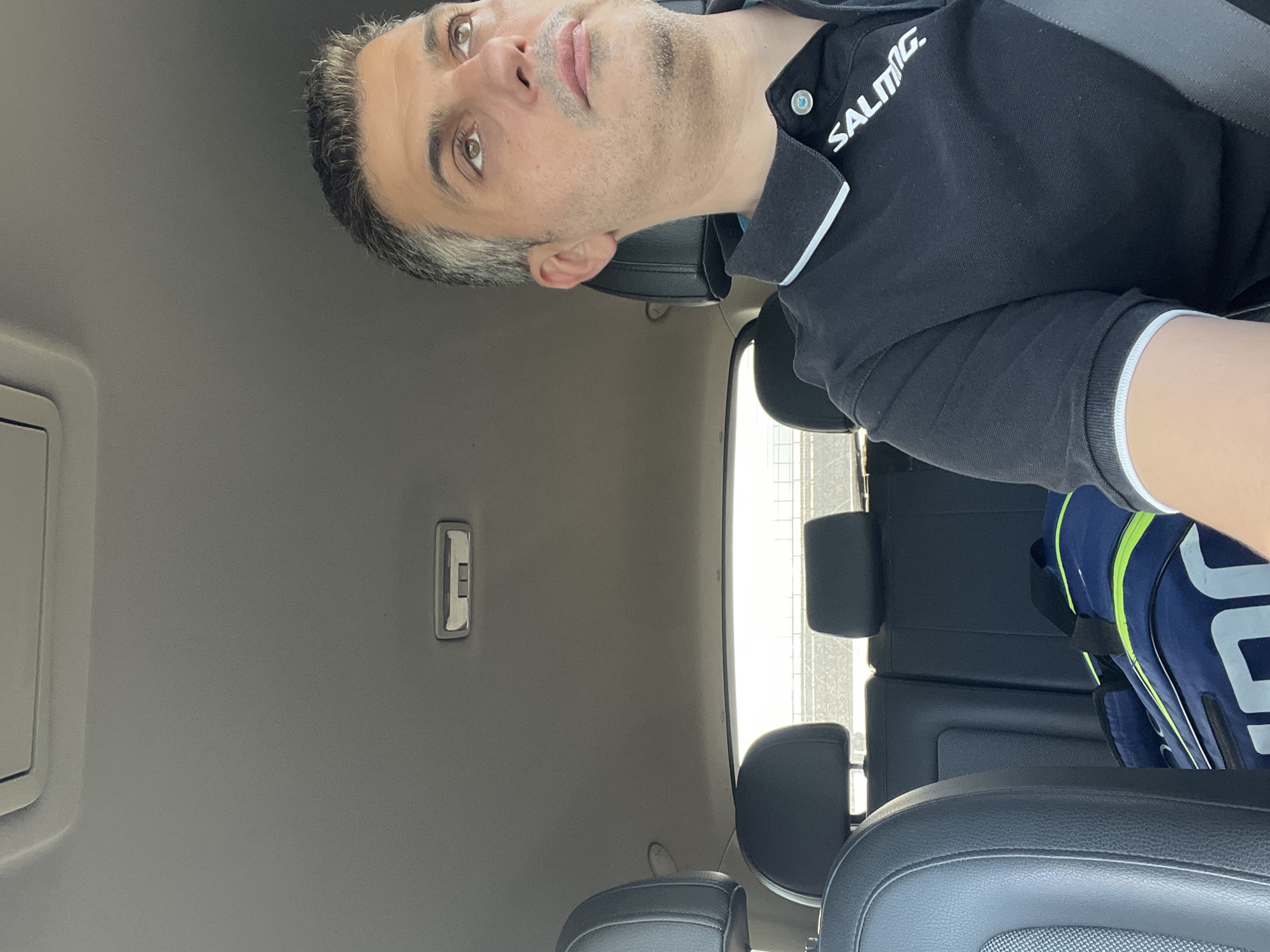 A medium shot captures a man with grey hair and a short beard driving a car. He is wearing a black polo shirt with white trim on the collar and sleeves, and the logo "SALMING" is visible on his shirt. The man is looking up and to his right. In the background, the car's interior is visible, including black leather seats and headrests. A blue and green duffel bag rests on the back seat. The scene appears to be during the daytime, with sunlight streaming in from the rear window. The specific location is not discernible from the image.