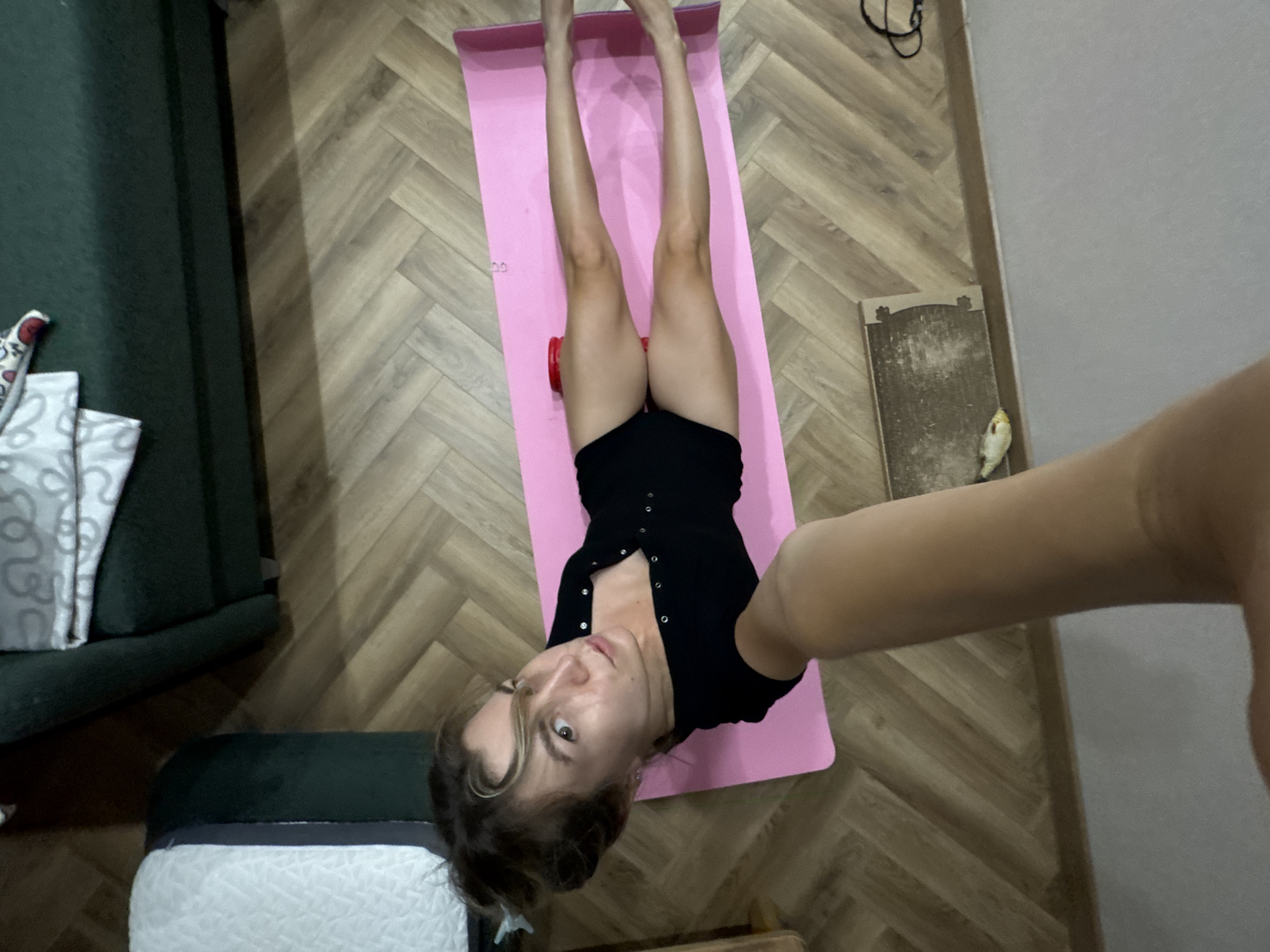 In an indoor setting, a woman is lying on a pink yoga mat on a wooden floor. She is wearing a black short-sleeved bodysuit. Her legs are extended, and her arms are raised, holding a camera for a selfie. A red circular object is placed under her right knee, and another under her left calf. Two couches, one dark green and the other partially visible and light-colored, are in the background. The floor is covered with a herringbone-patterned wood laminate. A small, light-colored bird is on the floor near the bottom edge of the image.