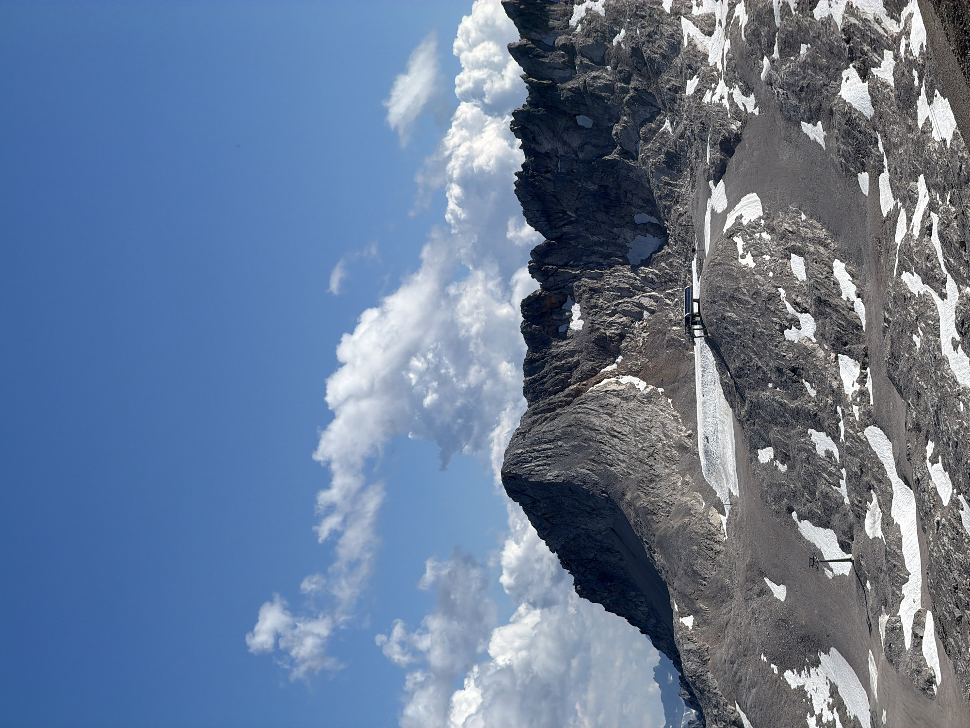 The image depicts a mountainous landscape under a bright blue sky with scattered white clouds. In the foreground, the rocky terrain is speckled with patches of snow. A lone ski lift pylon with a green support is visible on the left side of the frame. Further into the scene, a building with a dark roof structure is situated on a rocky outcrop, with a flat, open area in front of it. The background is dominated by a rugged mountain range with steep, jagged peaks, some of which show remnants of snow. The overall impression is of a high-altitude, possibly alpine, environment. This scene is likely the Zugspitze massif, the highest peak in Germany, located near Garmisch-Partenkirchen.