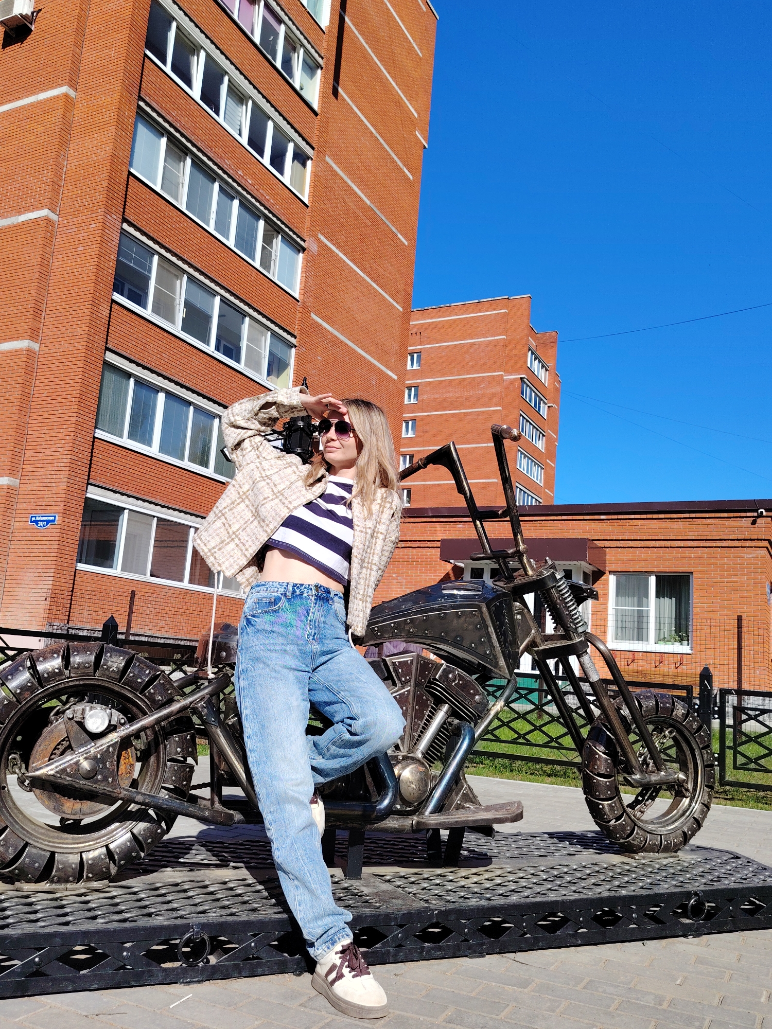 A medium, full shot captures a woman in a casual pose next to a custom-made metal motorcycle, with a brick building in the background.
The woman, fair-skinned with blonde hair, wears sunglasses, a striped navy and white crop top, light blue jeans, and a tweed jacket. Her left hand shades her eyes as she looks off-camera. Her left leg is propped up on the motorcycle.
The motorcycle is crafted from metal scraps and is permanently mounted on a black, grated platform.
The background features a large, modern, red brick apartment building with white-framed windows under a clear blue sky. A street sign reading "ul. Kalabushkina 24/1" is partially visible on the side of the apartment building.
The location is Chaikovsky, Russia.