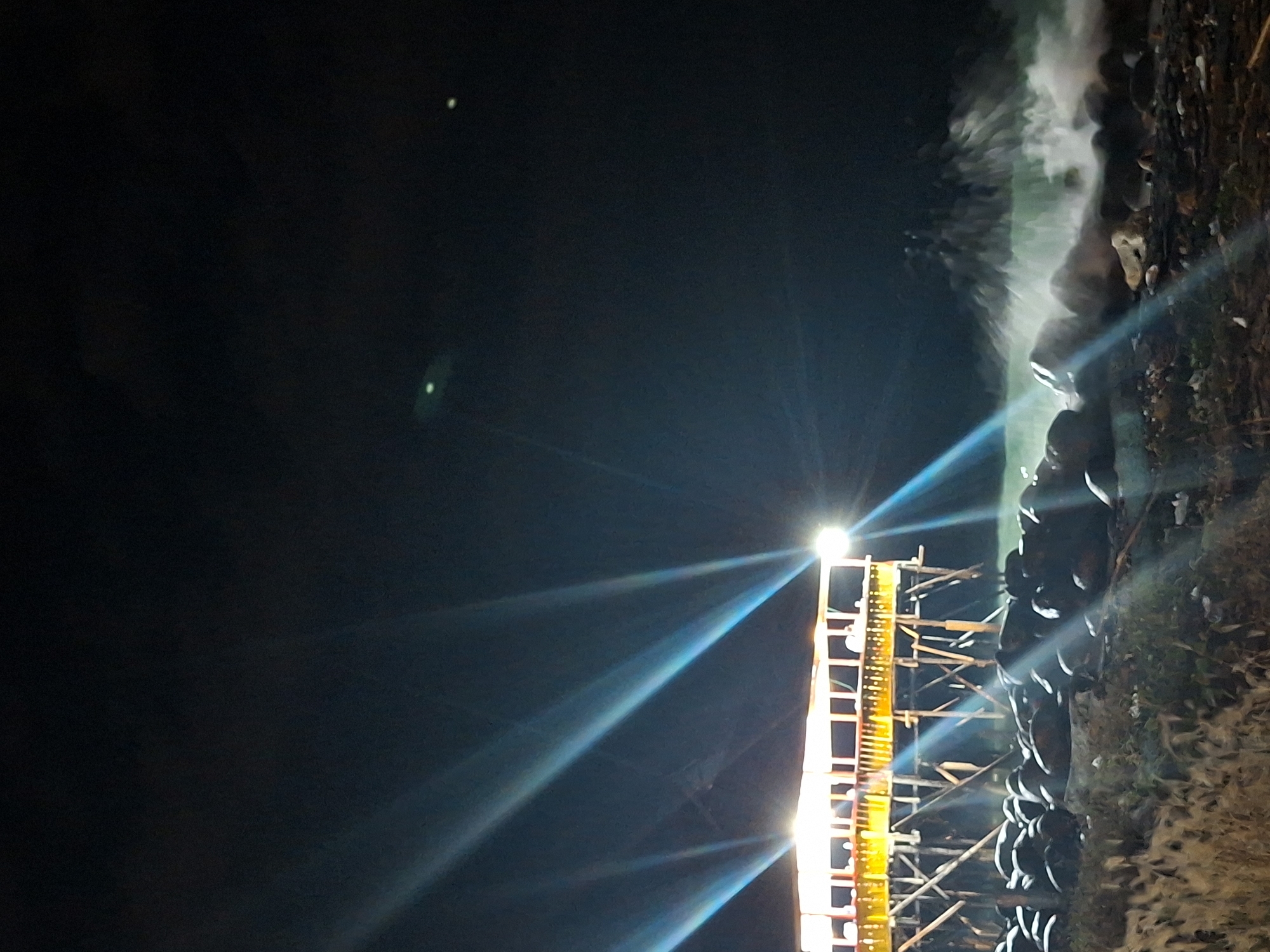 The image, oriented vertically, depicts a dark scene, seemingly at night, possibly in Lhokseumawe, Indonesia, given the request. A bright light source shines from a structure to the right, casting beams of light across the dark background. The structure appears to be a tall, possibly temporary, frame or scaffolding, constructed with yellow supports and horizontal elements. To the right of the scaffolding, is a dark, textured surface with dark rounded objects, potentially tires or stacked objects, and foliage. The overall impression is one of a construction site or outdoor event illuminated at night. A small bright spot is visible at the top left of the image in the dark sky.
