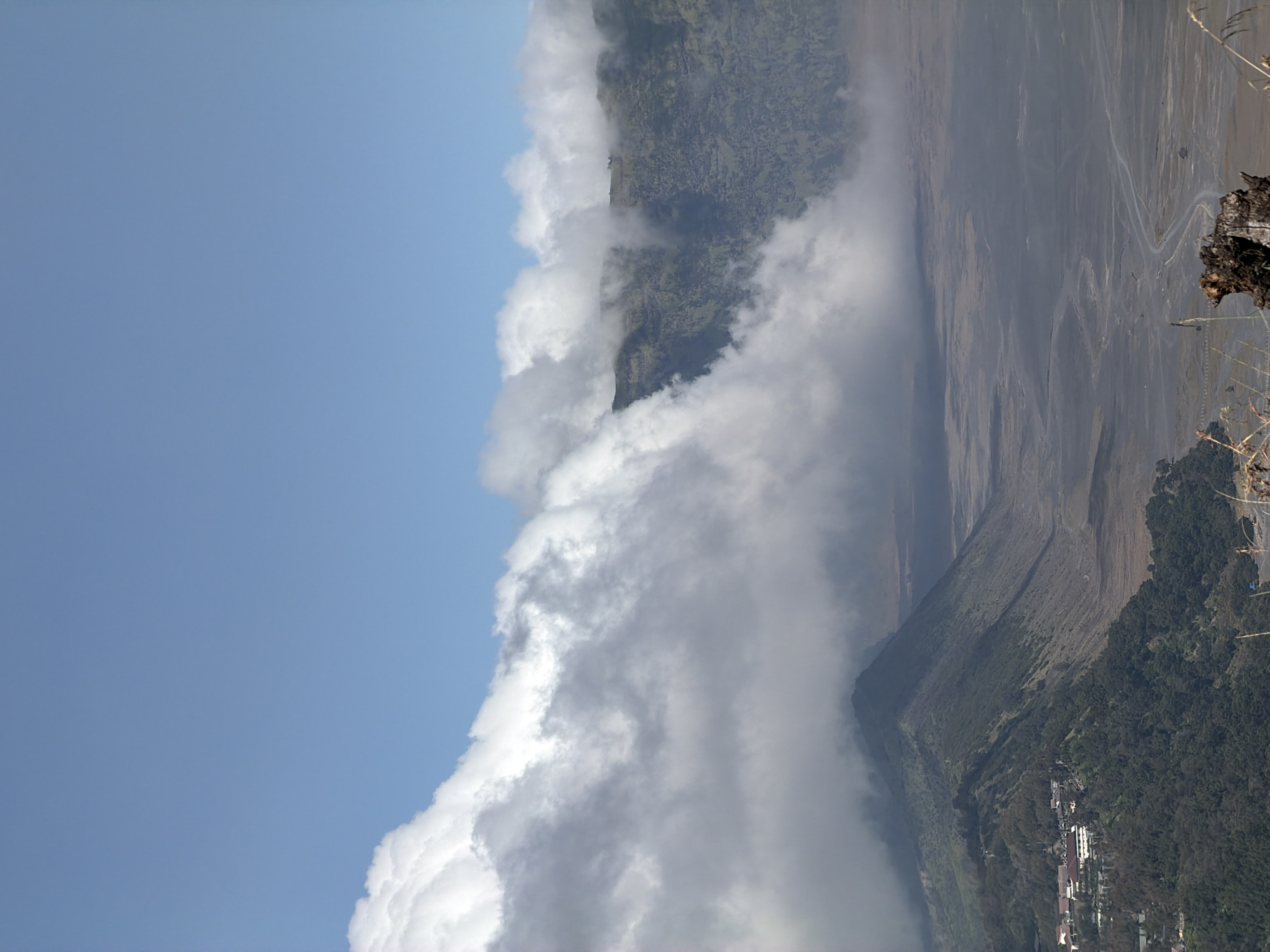 The image shows a panoramic view of a volcanic landscape with rolling hills, a vast plain, and a dramatic cloudy sky. In the foreground, a lush green hillside slopes down towards the right, dotted with trees and a cluster of buildings that appear to be part of a town or resort. A winding road is visible on the plain in the middle ground. The background features a barren, sandy plain stretching towards distant, hazy mountains. Clouds are a prominent element, partially obscuring the mountain peaks and creating a sense of depth and atmosphere. The sky above is a clear blue. The location is identified as Cemorolawang, Indonesia.